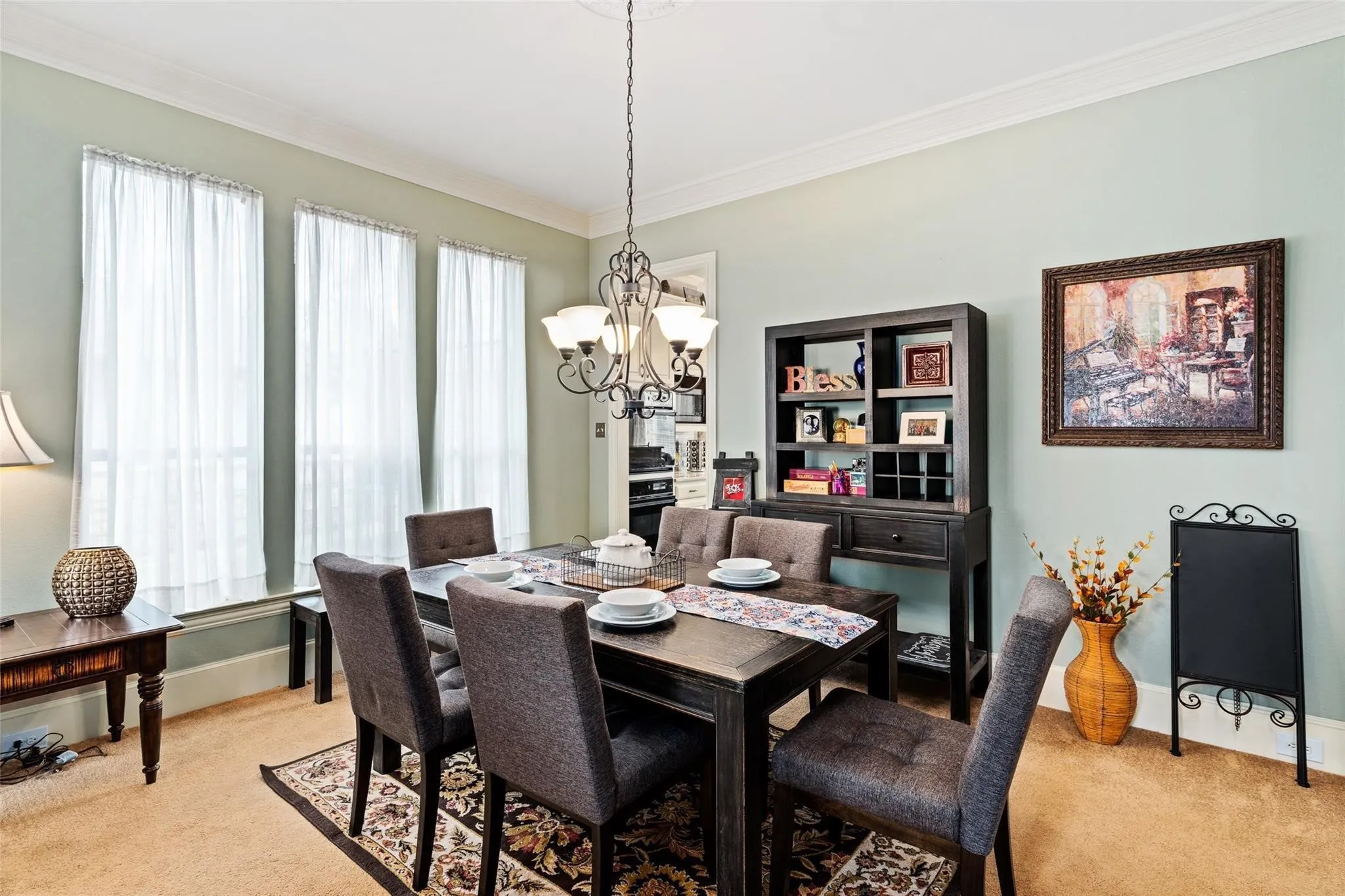 Dining area featuring a dark-finish dining table with upholstered chairs, three tall windows with sheer curtains, a decorative chandelier, and crown molding