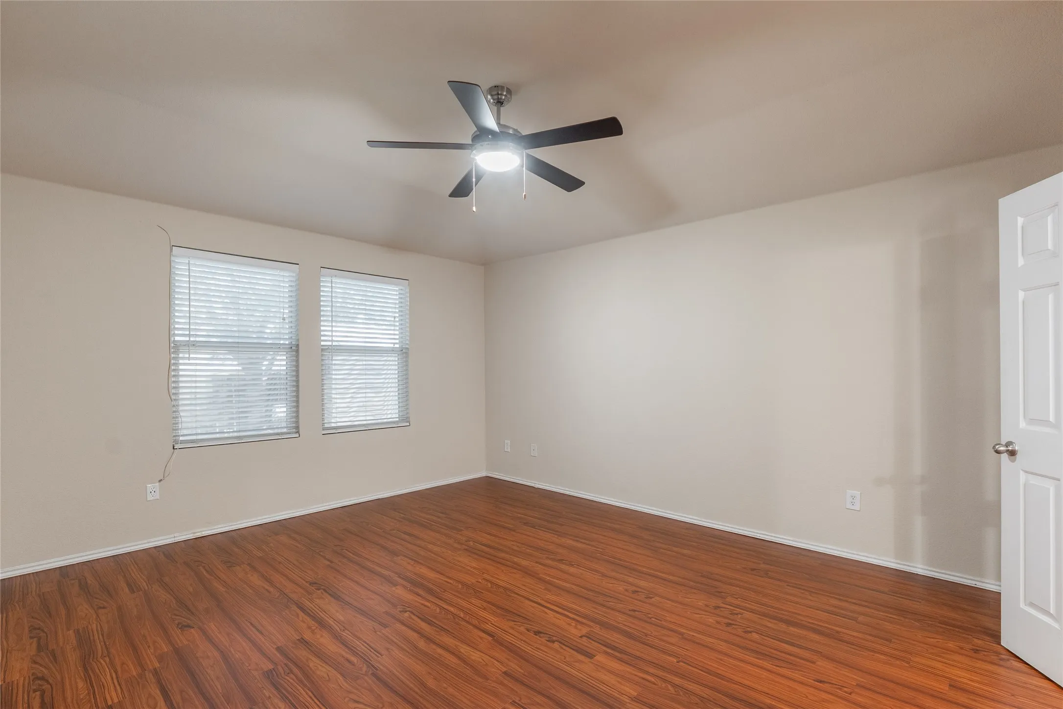Spacious room featuring wood-finish flooring, light neutral walls, two windows with blinds, and a ceiling fan with integrated lighting