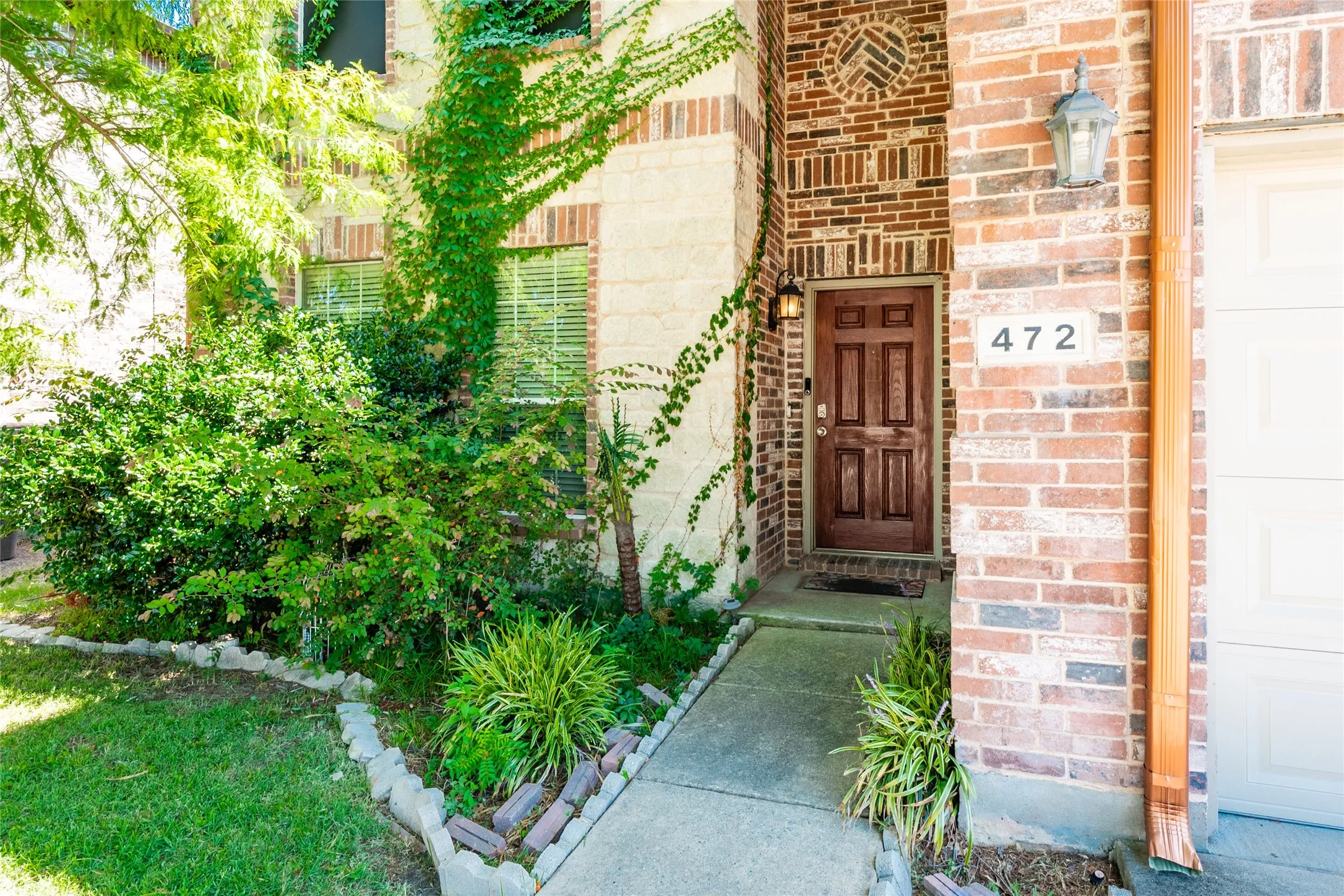 Brick and stone facade featuring a paneled wood-finish entry door, concrete walkway, and established landscaping