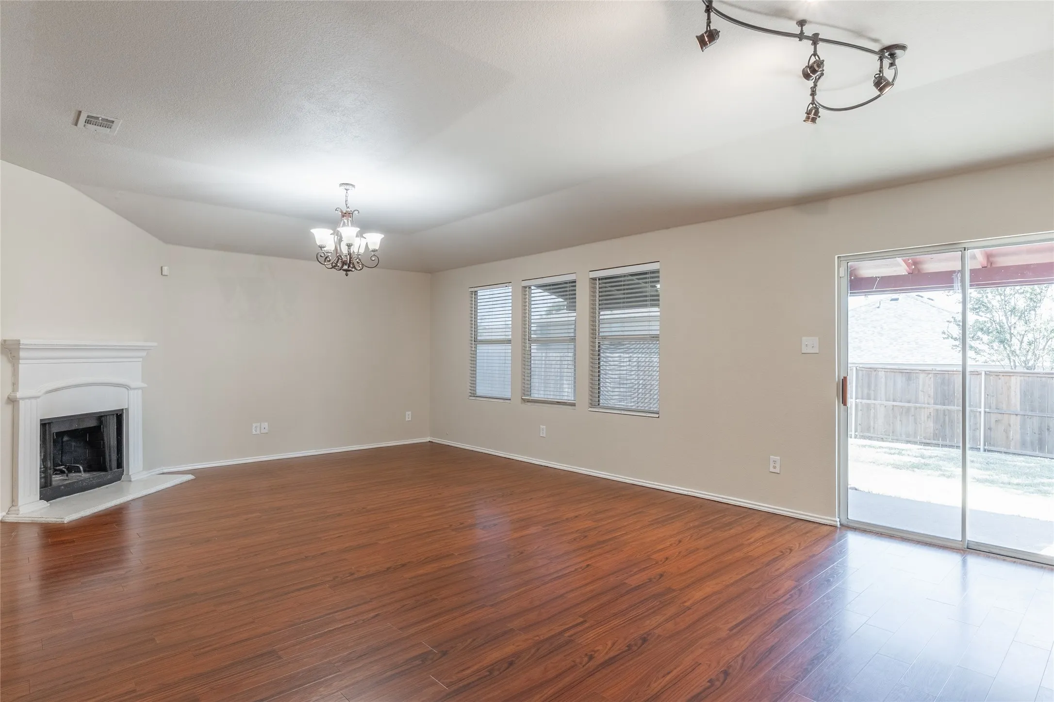 Spacious living area featuring wood-finish flooring, a corner fireplace with white mantel, a multi-light chandelier, three windows with blinds, and a sliding glass door opening to a patio
