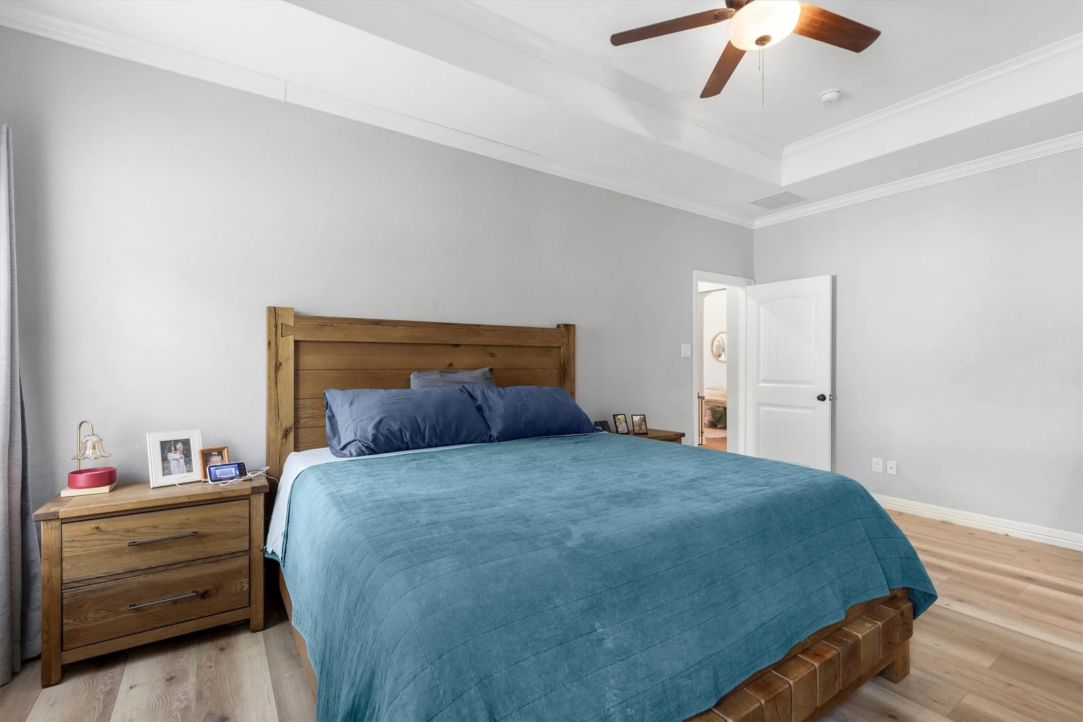 Spacious bedroom featuring light wood-finish flooring, a tray ceiling with crown molding, and a built-in ceiling fan with integrated lighting