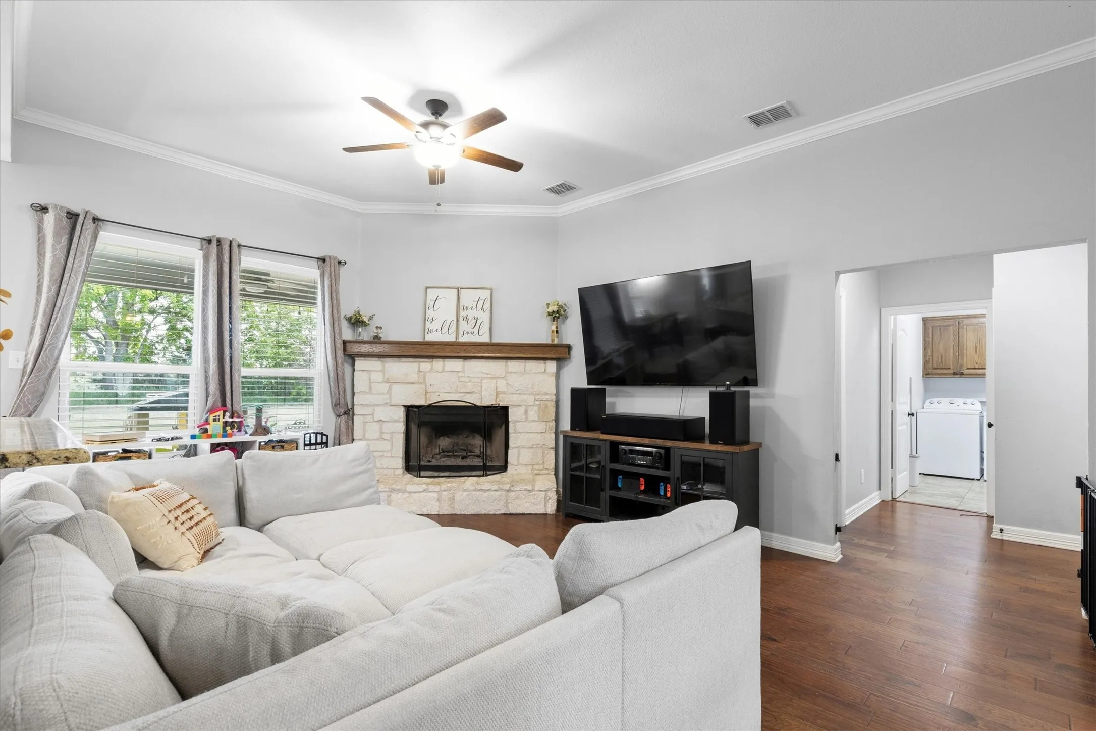 Spacious living area featuring a stone-clad fireplace with a wood mantle, ceiling fan, crown molding, and wood-finish flooring