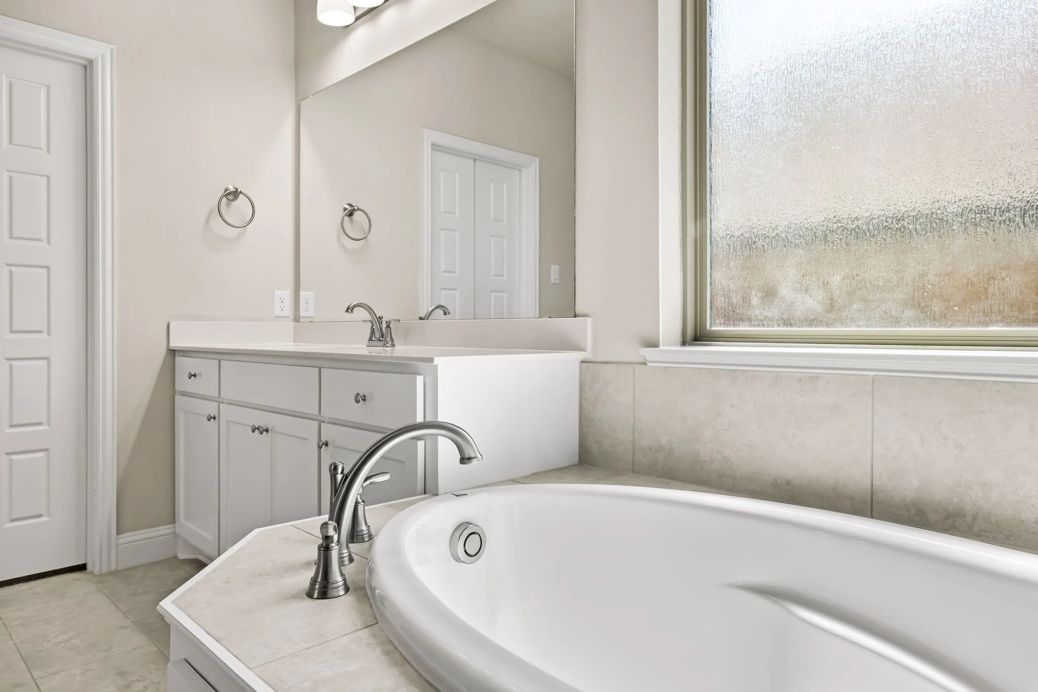 Soaking tub with tile surround, frosted window, dual vanity with white cabinetry, polished chrome fixtures, and a large wall mirror