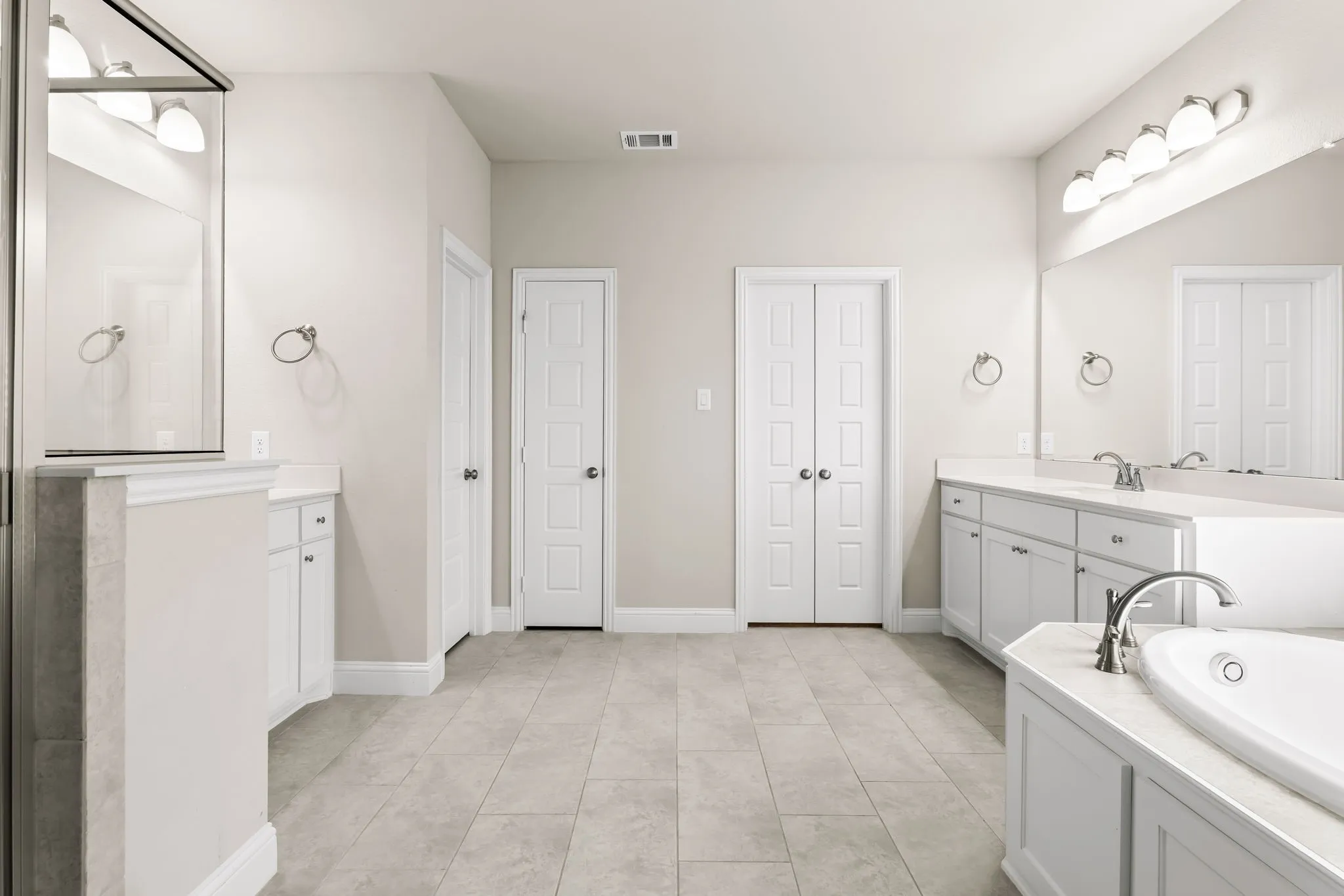 Spacious bathroom featuring light neutral wall paint, tiled flooring, dual vanities with white cabinetry, built-in bathtub, and panel doors