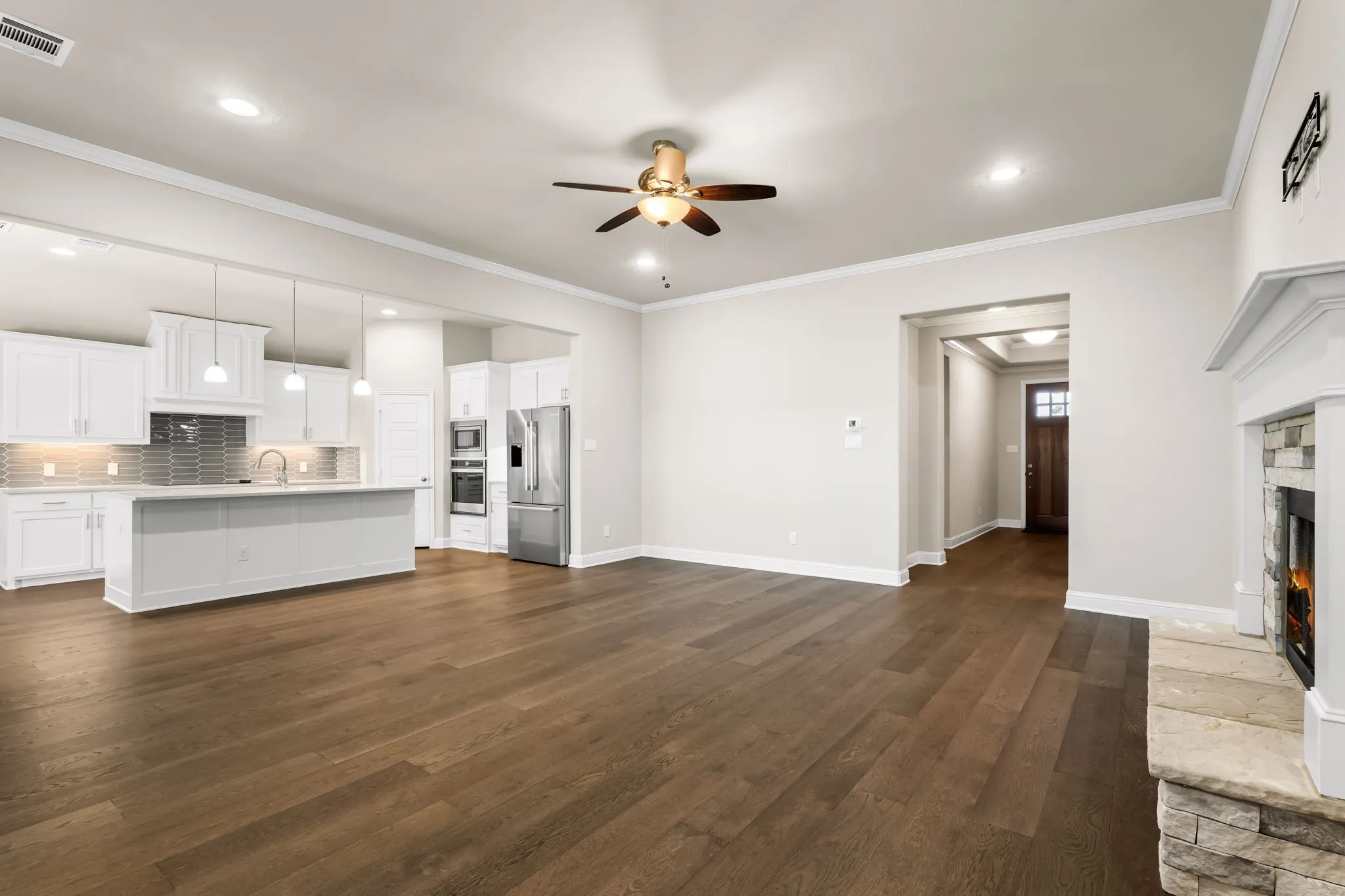 Open-concept living area featuring wood-finish flooring, a stone-surround fireplace with a white mantel, and a kitchen with white cabinetry, stainless steel appliances, and a central island