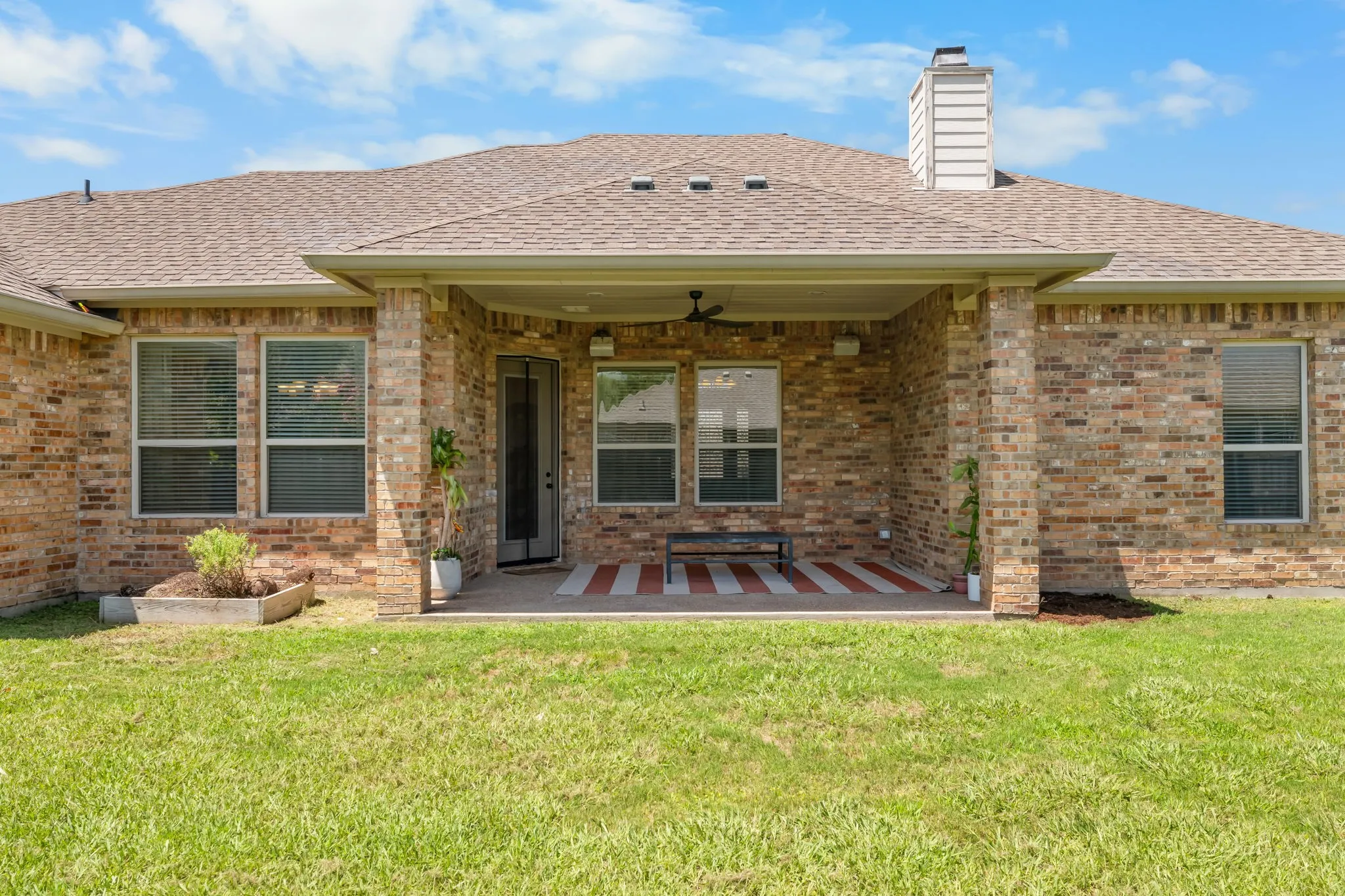 Spacious backyard featuring a covered patio with a ceiling fan, brick exterior, multiple windows, and a raised planter box