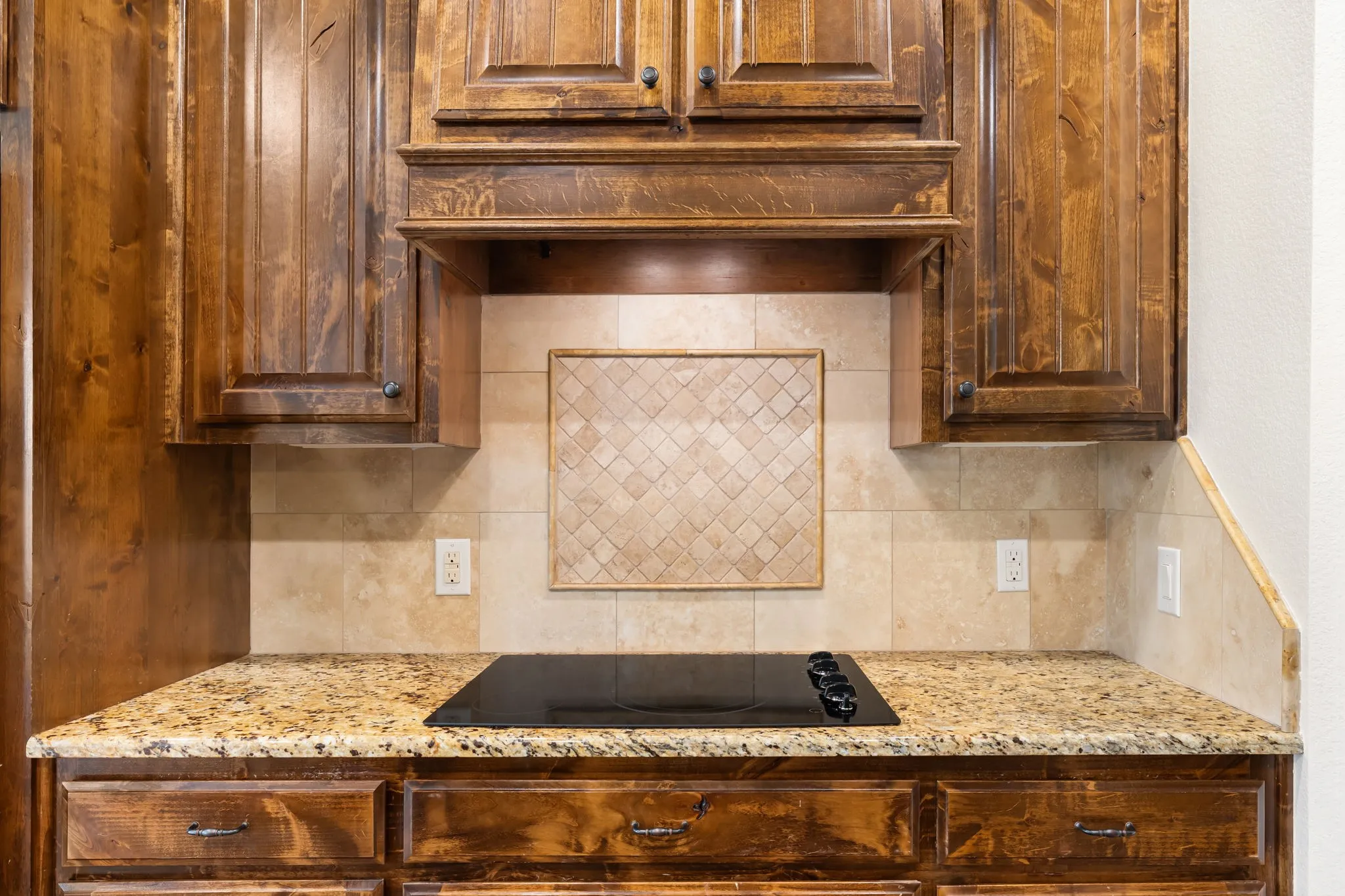 Kitchen area featuring rich wood cabinetry, patterned stone tile backsplash, a glass-top electric cooktop, and a speckled stone countertop