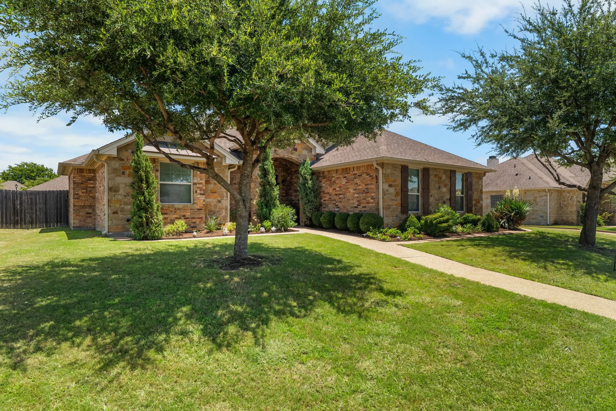 Brick and stone facade residence featuring a front yard, a paved walkway, mature trees, and established landscaping