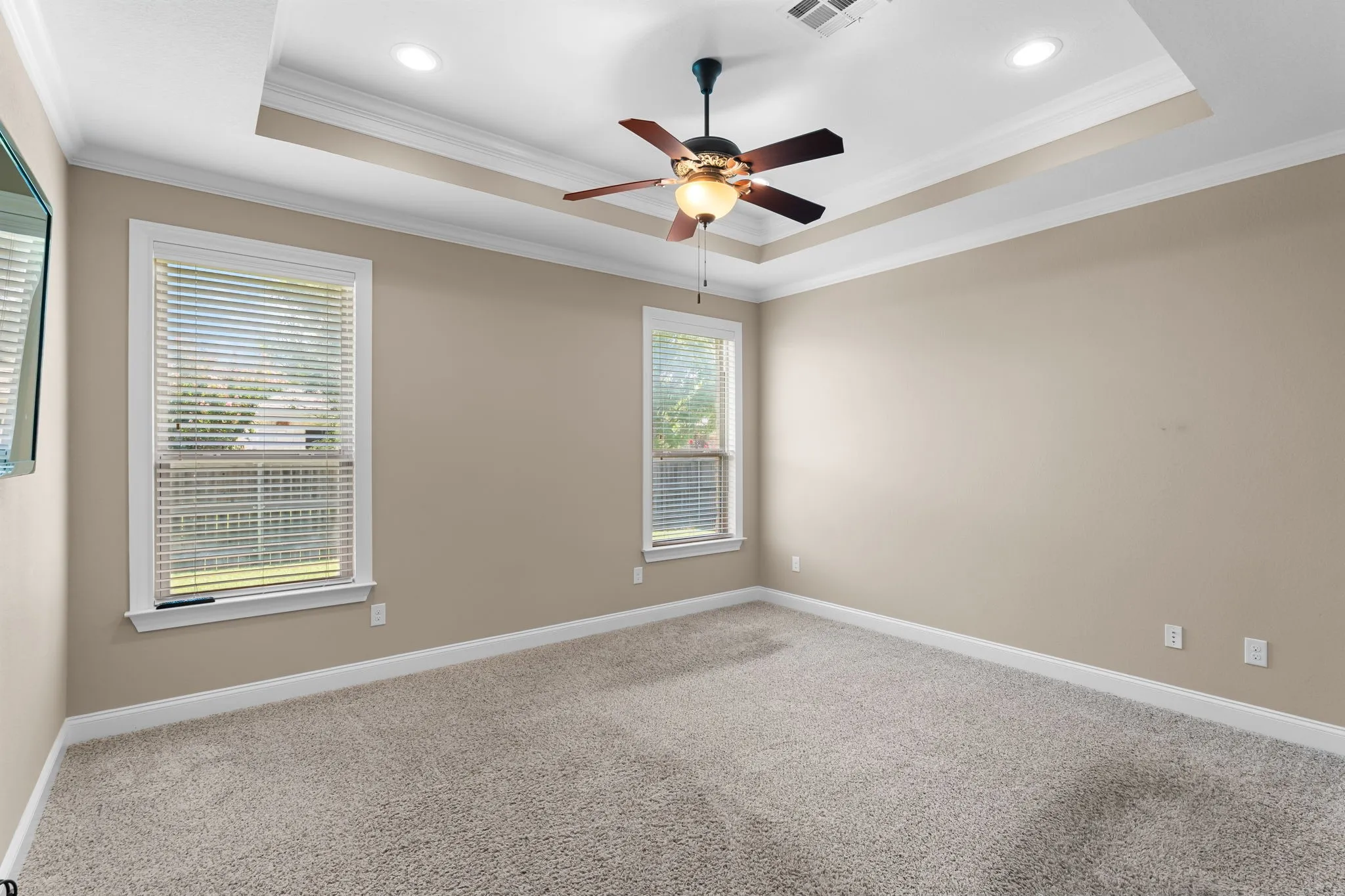 Recessed ceiling with crown molding, ceiling fan with light fixture, two windows with blinds, neutral wall paint, and carpet flooring