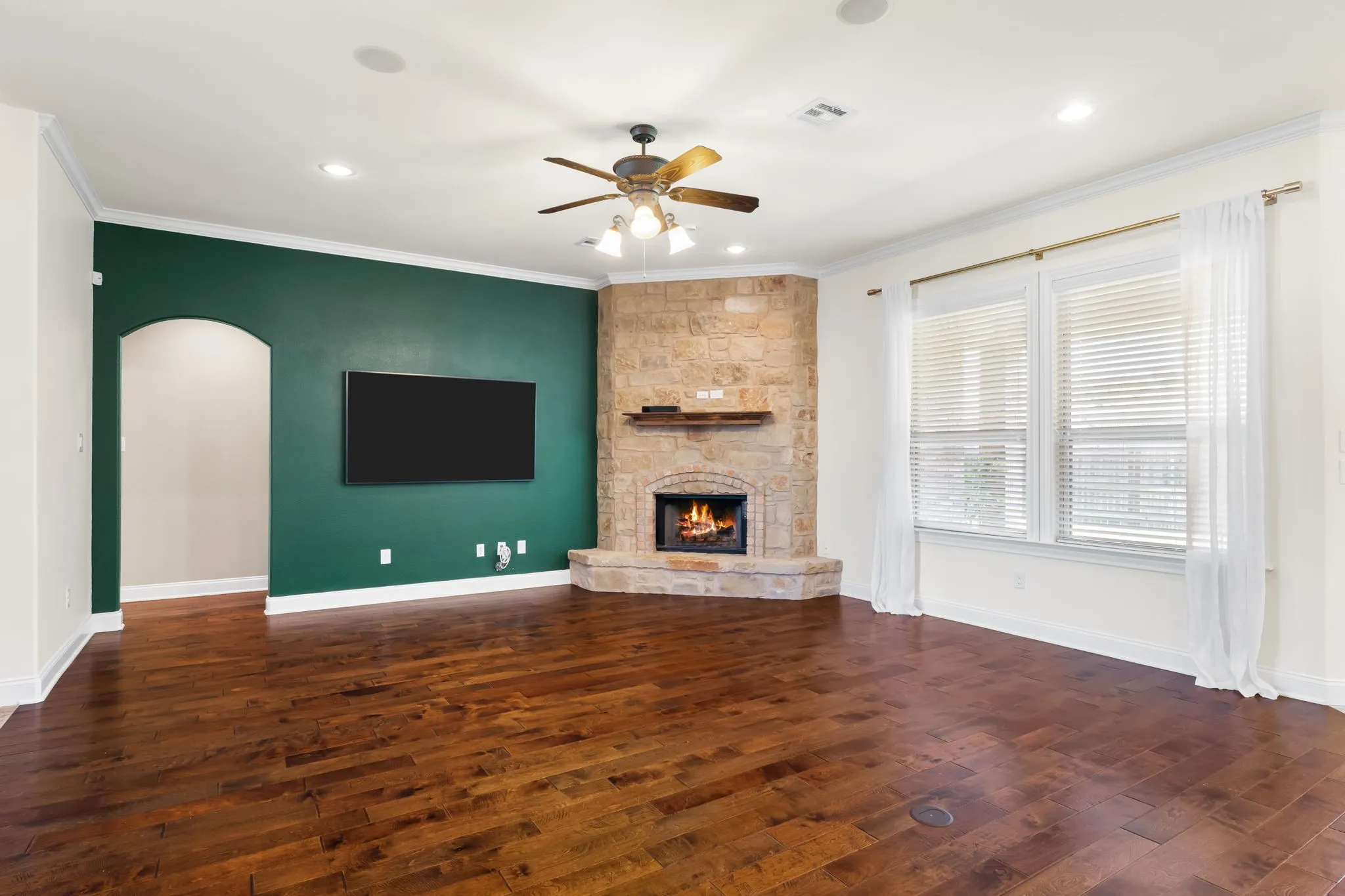 Spacious living area featuring wood-finish flooring, a corner stone fireplace, and a ceiling fan with integrated lighting
