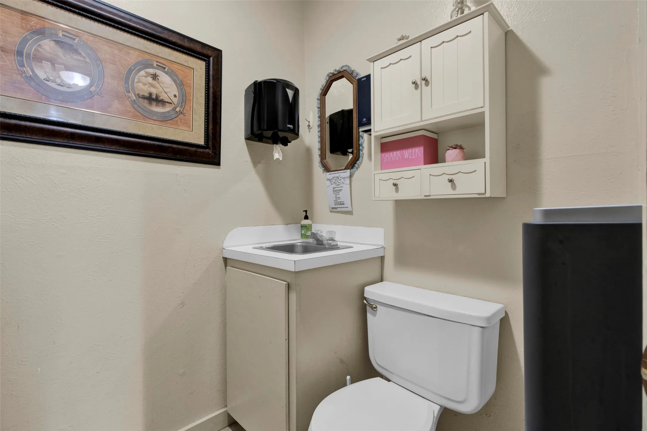 Compact bathroom featuring a white toilet, a vanity with an integrated sink and white countertop, and a wall-mounted cabinet with two drawers and shelving