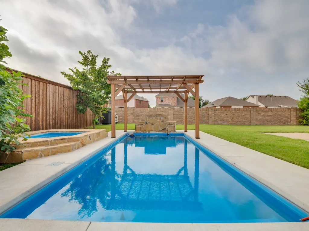 Backyard oasis featuring a rectangular swimming pool with a stone waterfall feature, an elevated hot tub with stone surround, a wooden pergola, and concrete decking