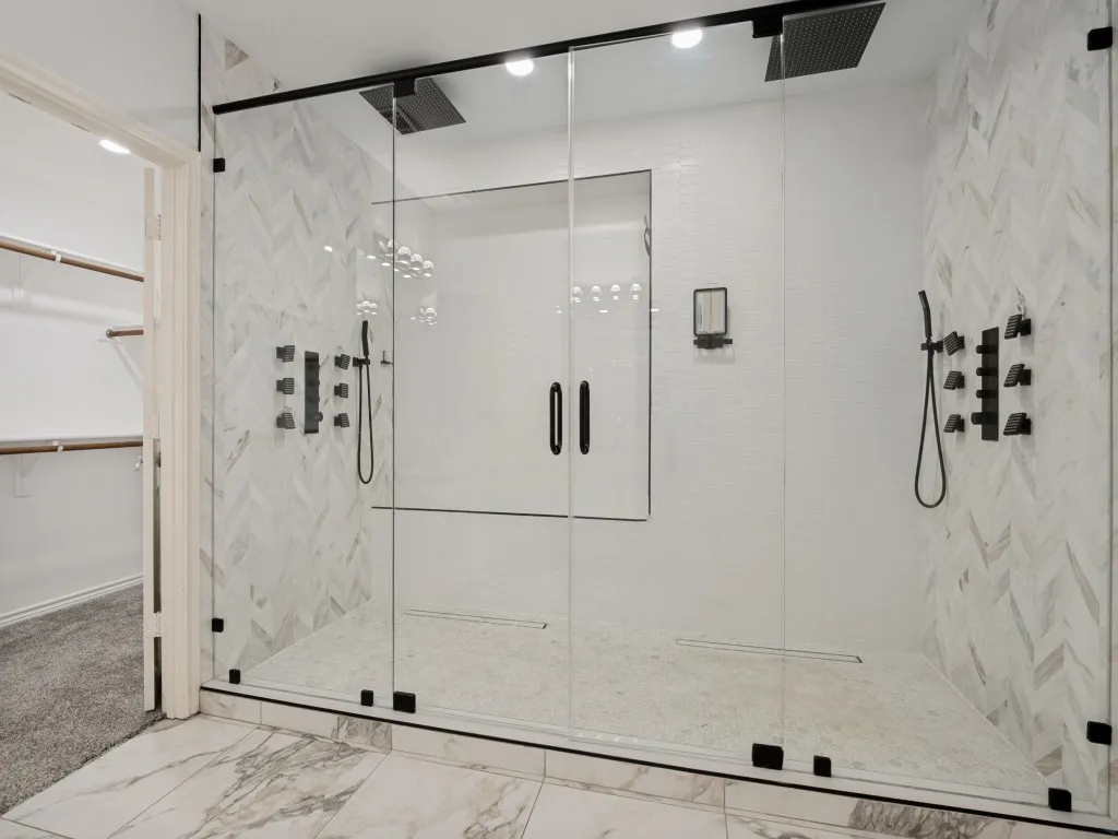 Spacious walk-in shower featuring clear glass enclosure with matte black hardware, dual overhead rain shower heads, multiple body spray jets, and white herringbone tile surround