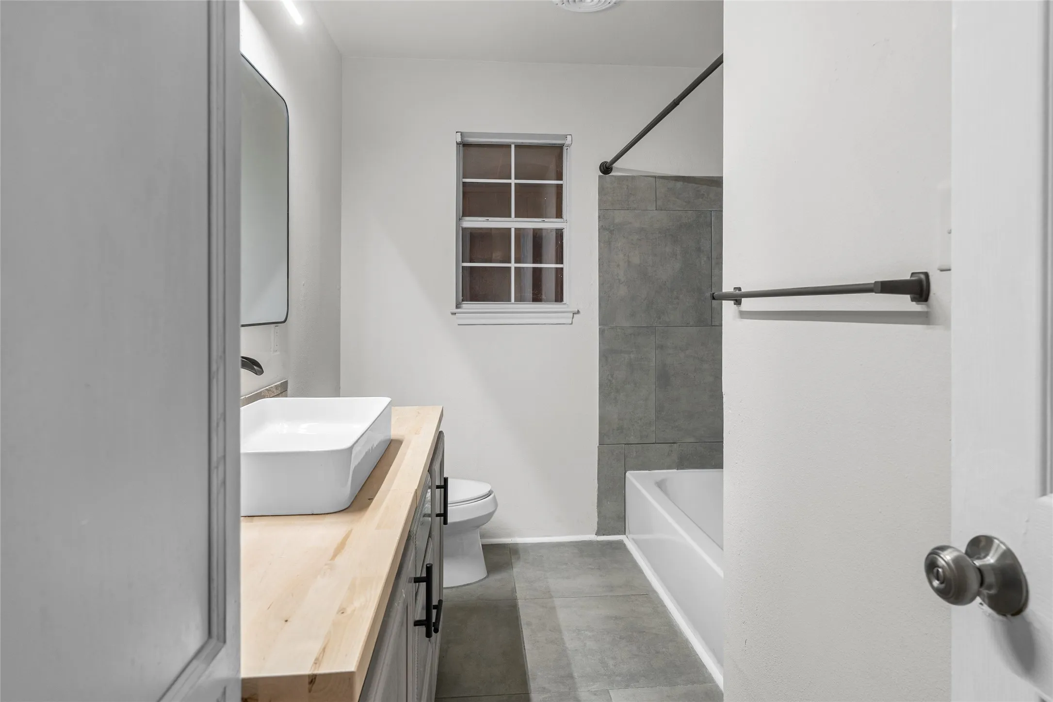 Bathroom featuring a rectangular vessel sink set on a wood-finish countertop vanity with black hardware, a rectangular vanity mirror, and a bathtub with gray subway tile surround