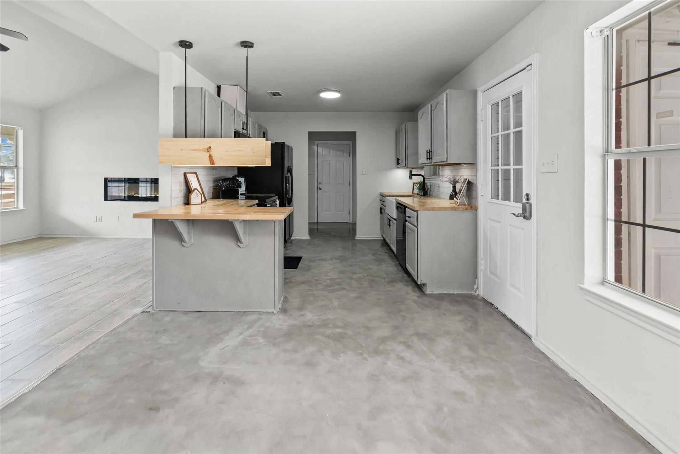 Open-concept kitchen featuring concrete flooring, light gray cabinetry, wood-finish countertops, and a built-in electric fireplace in the living area