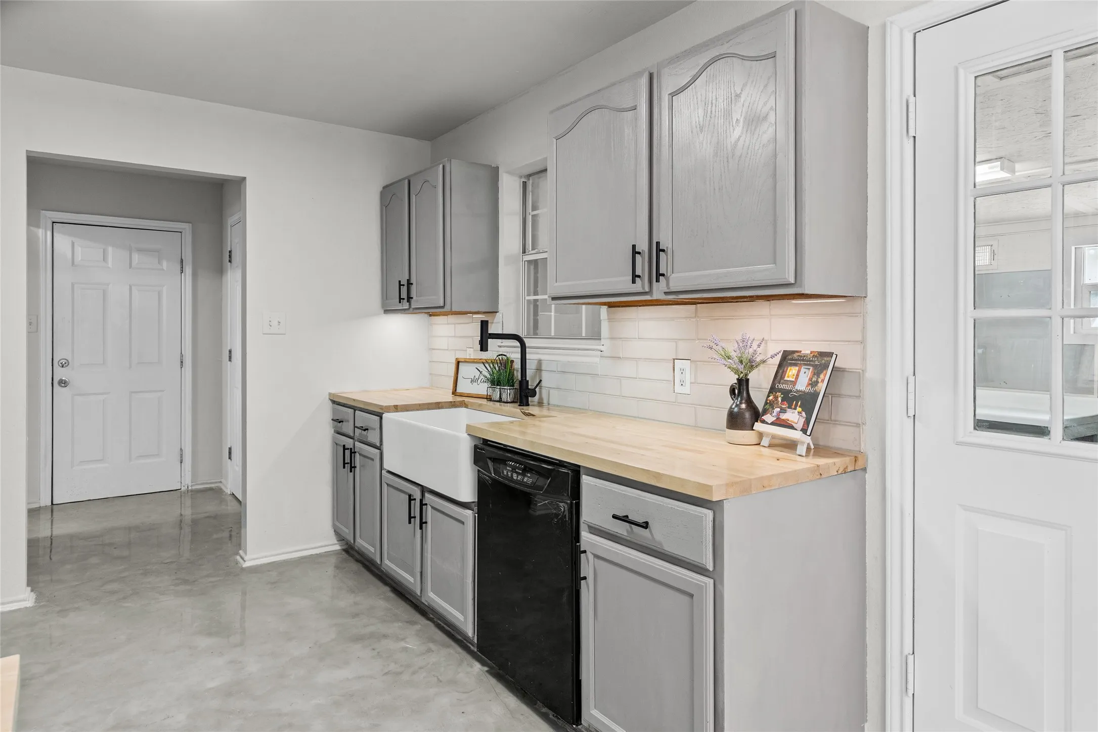 Kitchen area featuring gray cabinetry with black hardware, butcher block countertops, a white farmhouse sink with a black gooseneck faucet, a white subway tile backsplash, and a built-in dishwasher