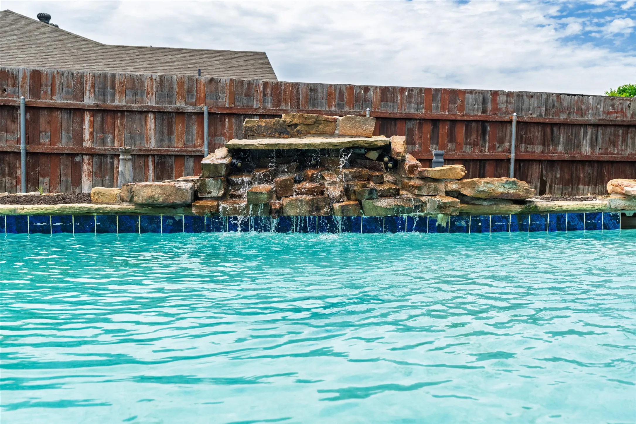 Swimming pool featuring a rock waterfall, blue waterline tiles, and a surrounding privacy fence