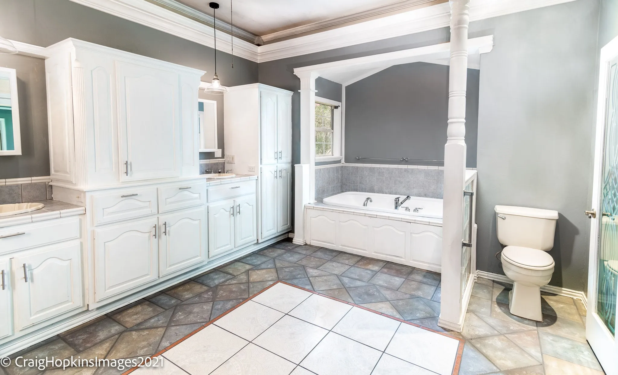Spacious bathroom featuring an oversized soaking tub, dual vanity areas, extensive white cabinetry, and mixed-tile flooring