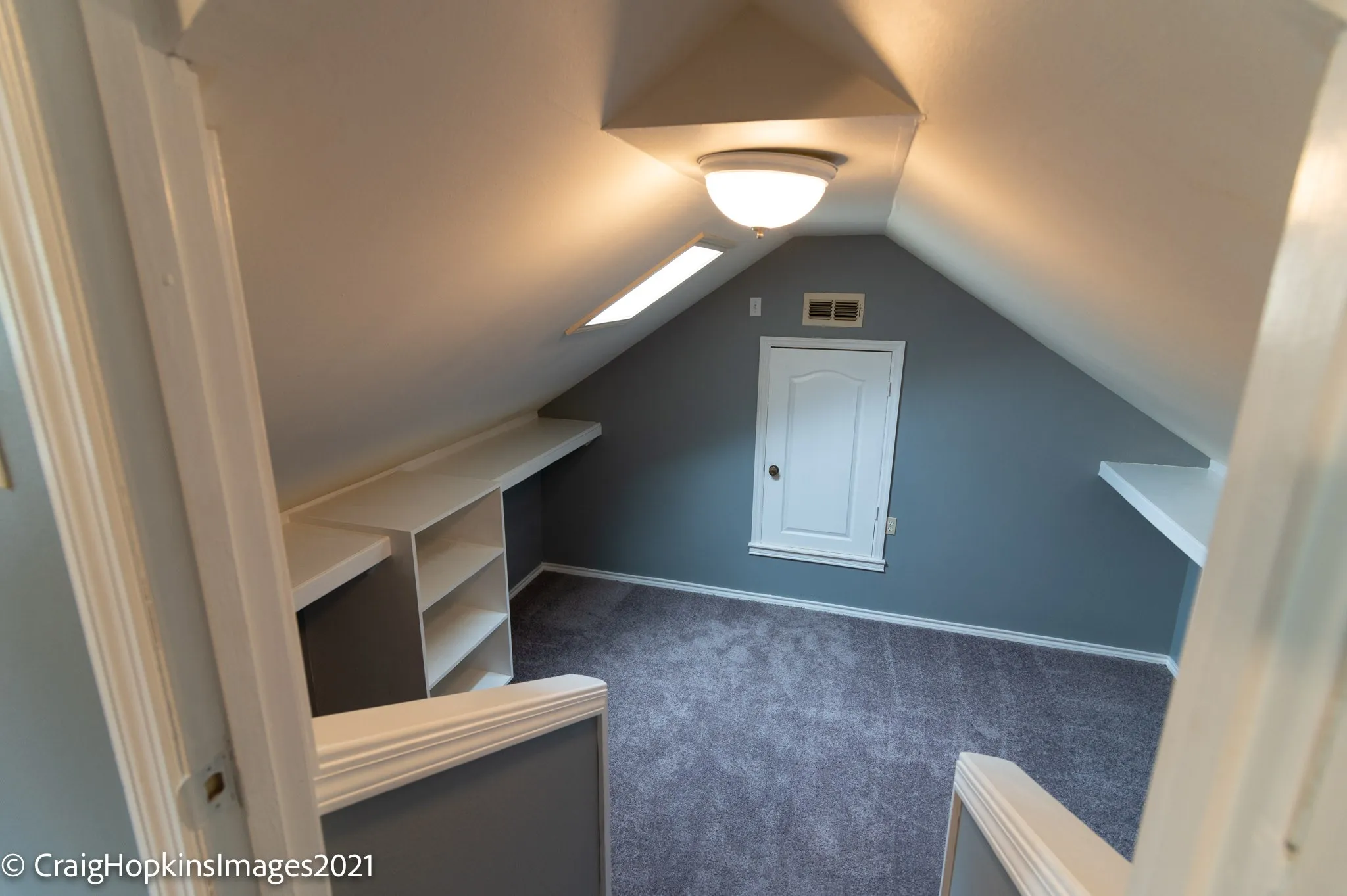 Carpeted attic space featuring sloped ceilings, two skylight-style windows, and a built-in shelving unit