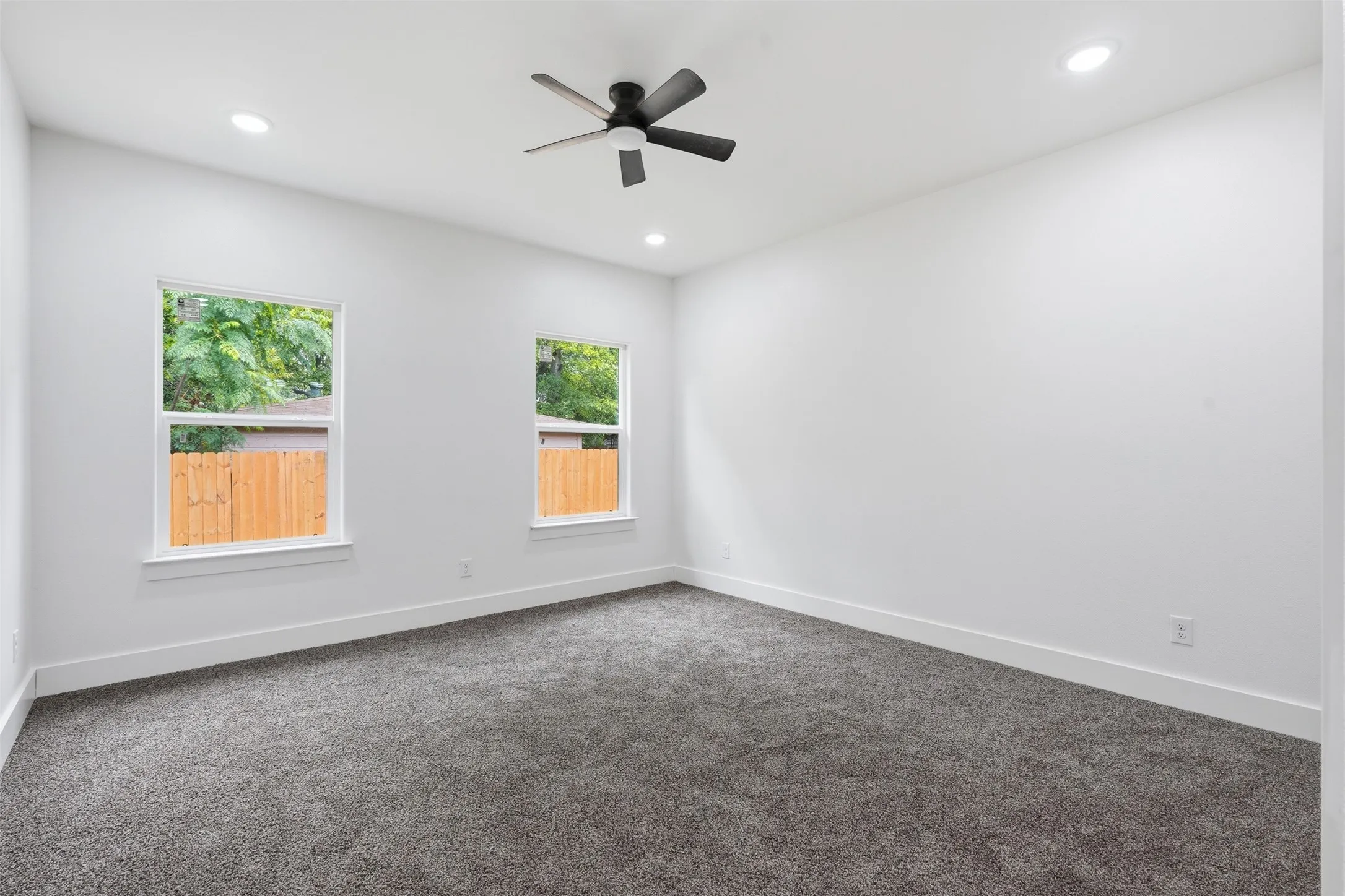 Spacious room featuring plush gray carpeting, crisp white walls, two windows with white trim, recessed lighting, and a ceiling fan with matte black blades