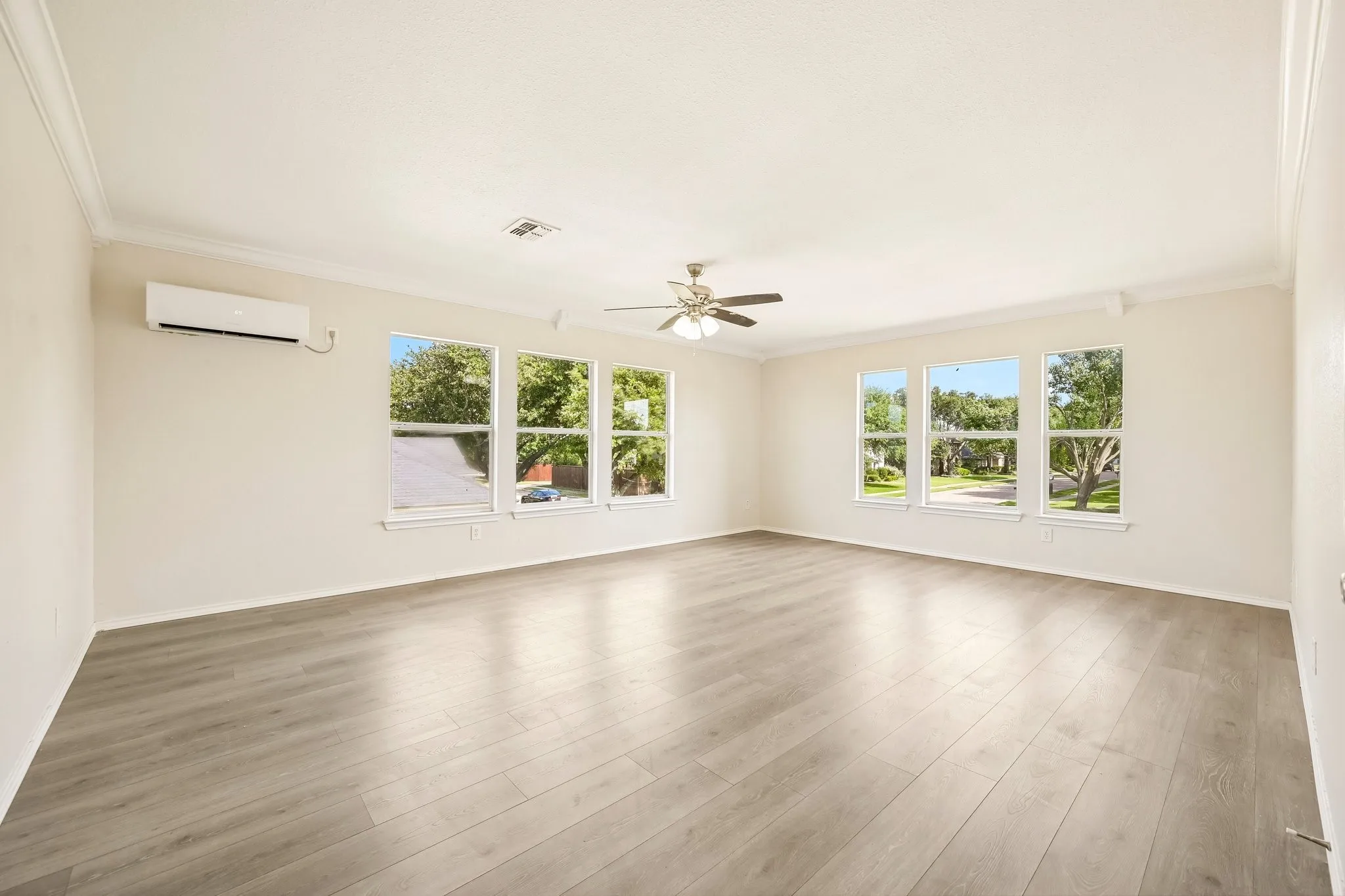 Expansive room featuring light wood-finish flooring, multiple windows, crown molding, a ceiling fan with integrated lighting, and a wall-mounted mini-split AC unit