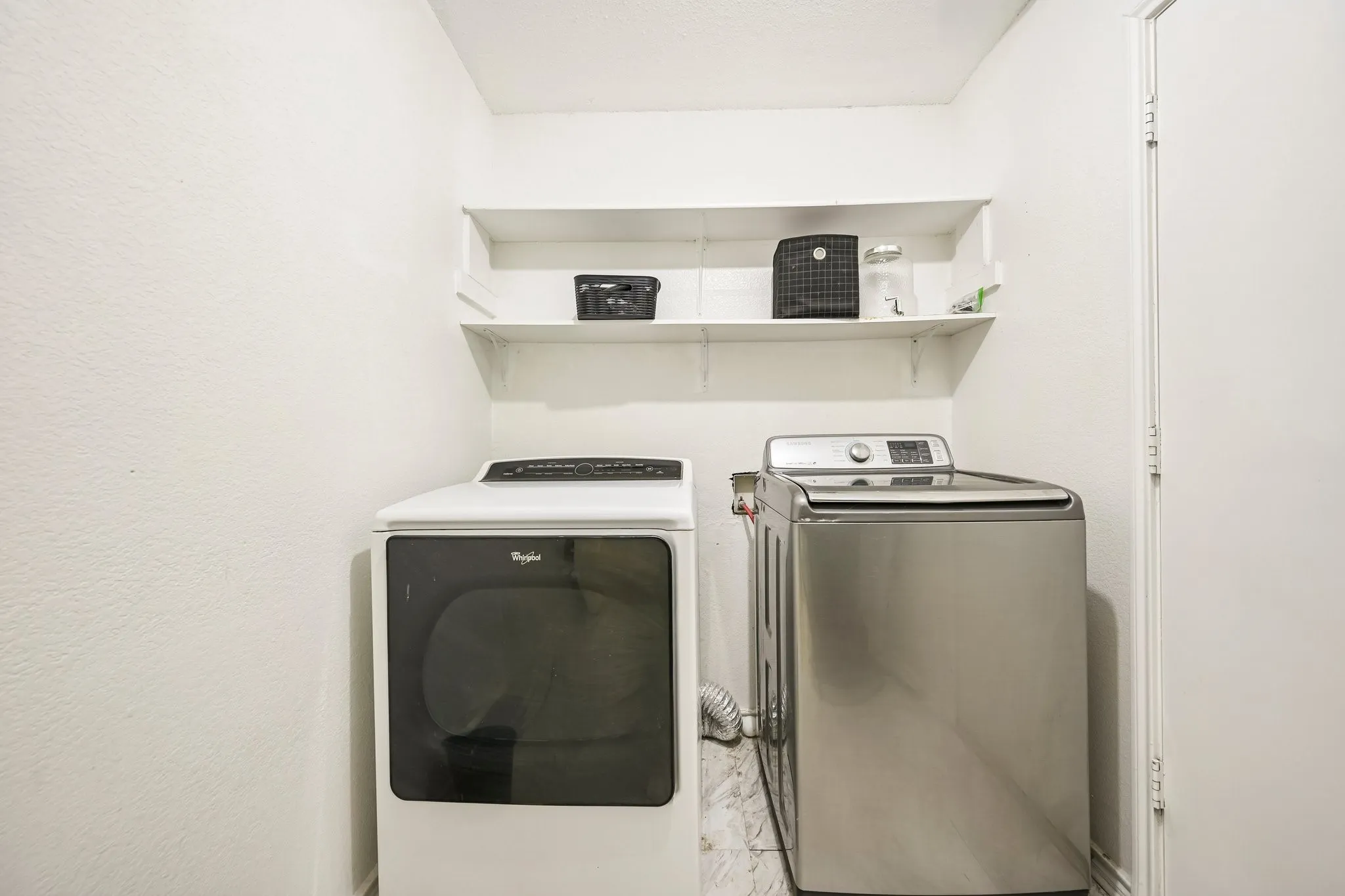 Dedicated laundry area featuring a white front-load dryer, a stainless steel top-load washing machine, two white wall-mounted shelves, and marble-look floor tile
