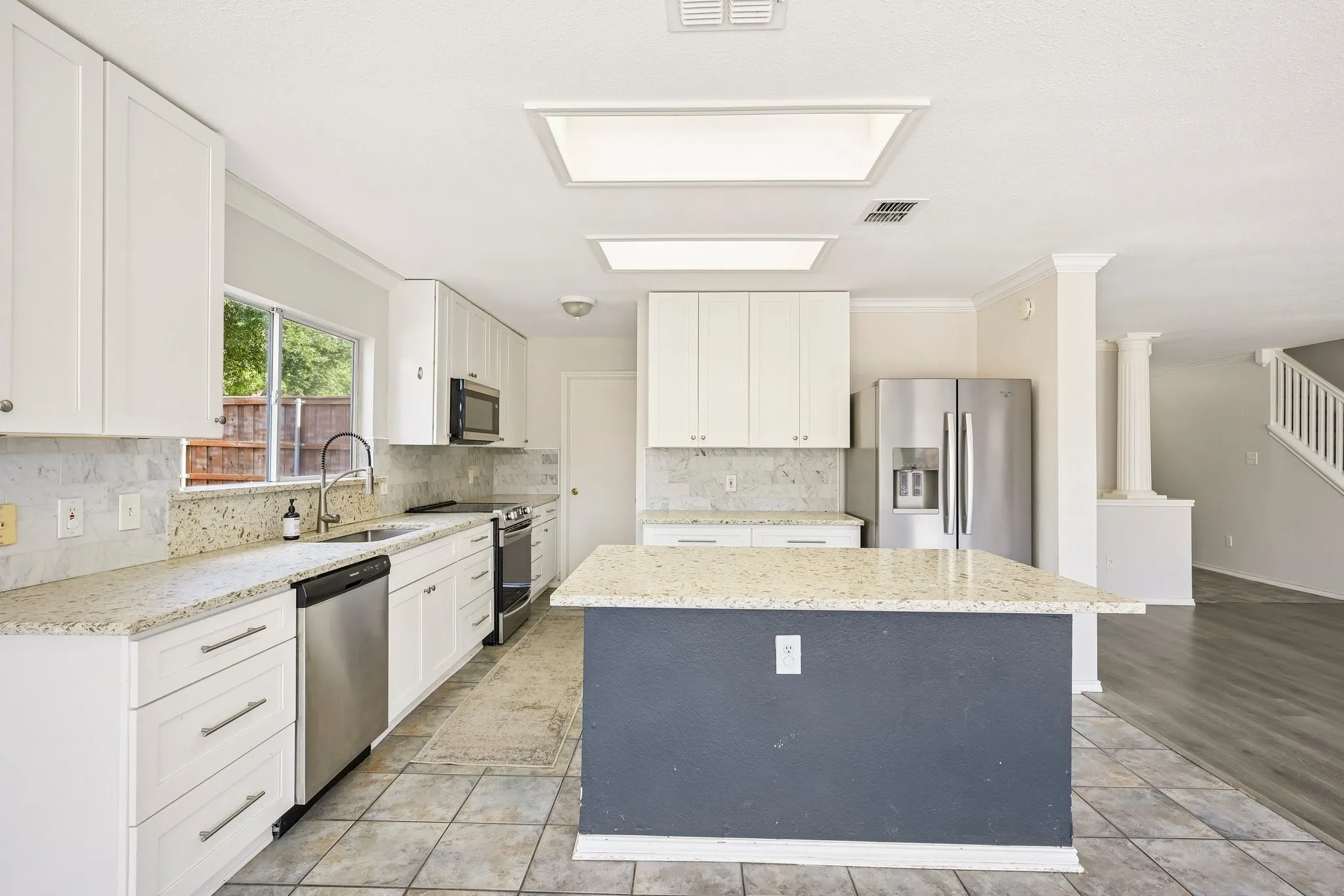 Bright kitchen featuring a central island with a granite countertop, white shaker-style cabinetry, stainless steel appliances, and tile flooring