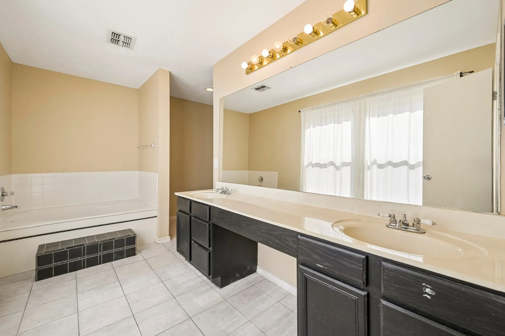 Bathroom featuring a built-in jetted tub with tile surround, a spacious vanity with dual sinks, a large wall-mounted mirror, and tile flooring