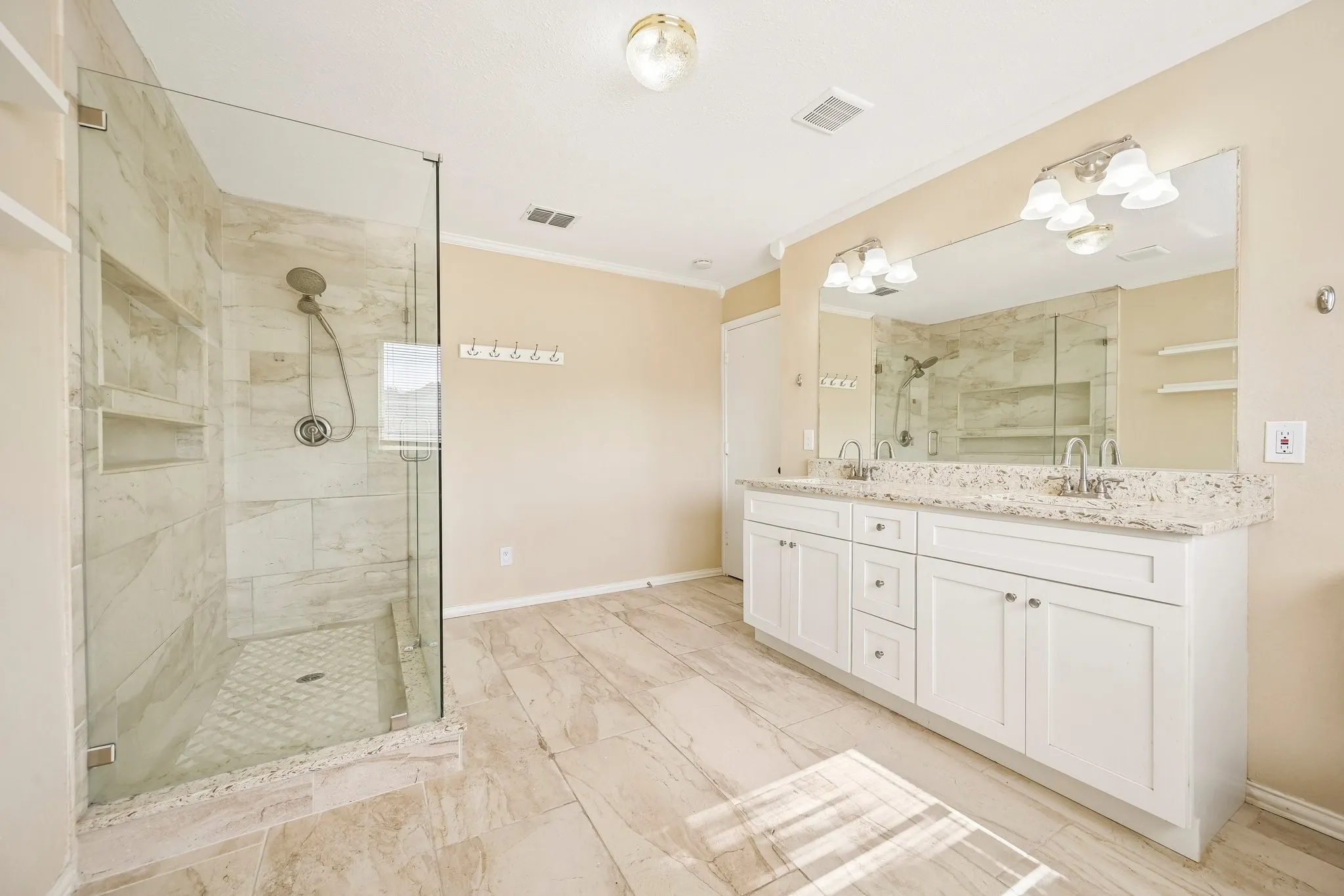 Spacious bathroom featuring a frameless glass shower enclosure with tiled walls and built-in niches, dual vanity with white cabinetry, granite countertops, and a large mirror