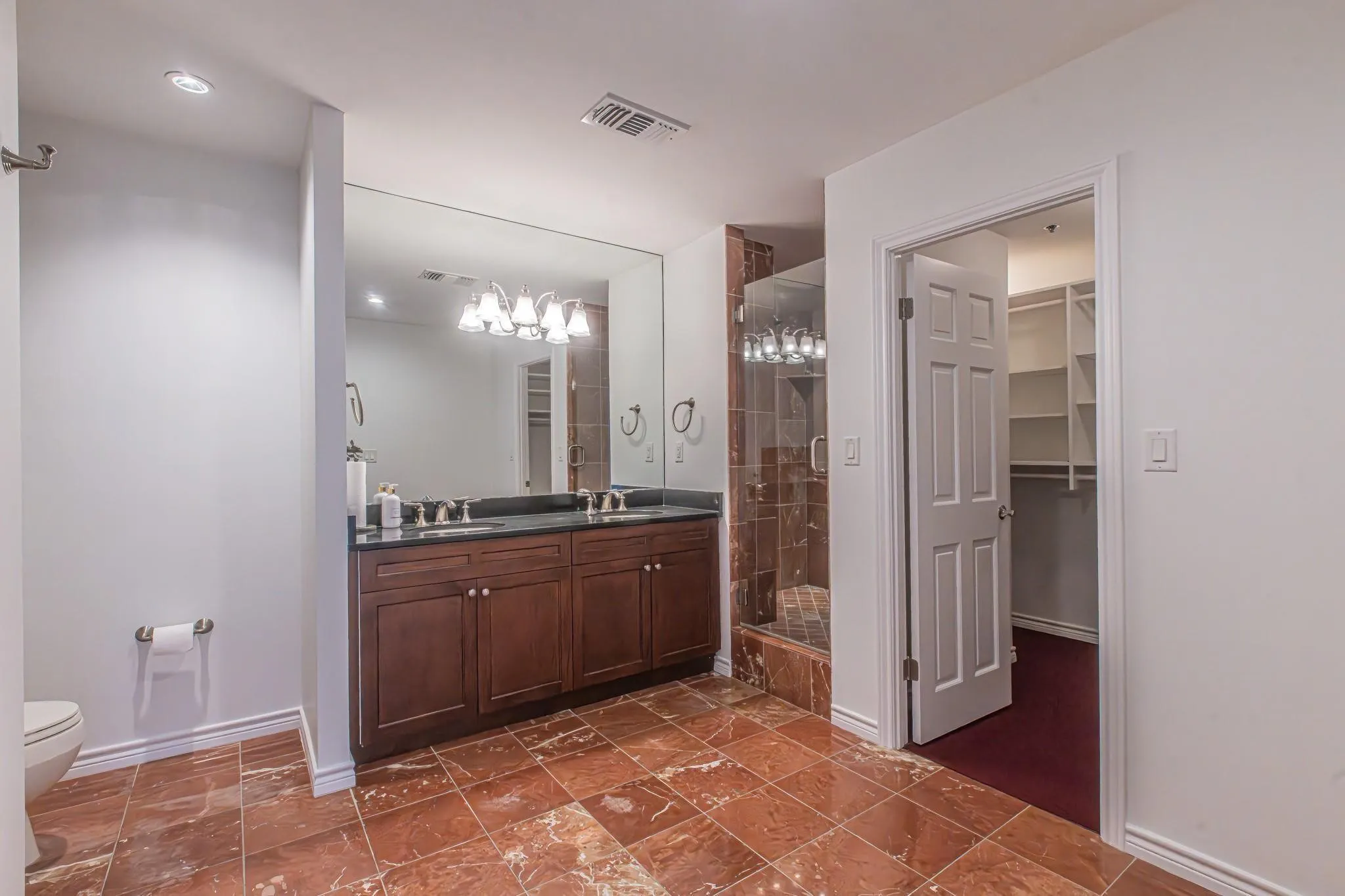 Bathroom featuring a double vanity with dark cabinetry and countertop, an expansive wall mirror, a glass-enclosed shower with matching tile, and red-toned floor tiles