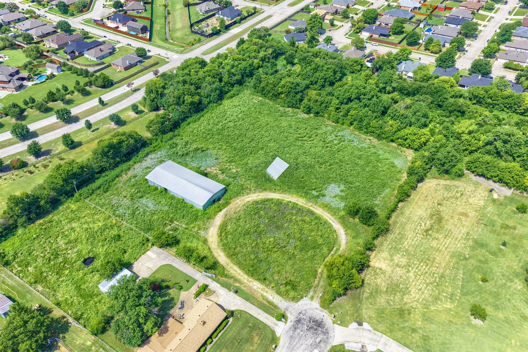 Expansive green acreage featuring a large metal building, a smaller utility structure, a winding dirt path, and surrounding mature trees