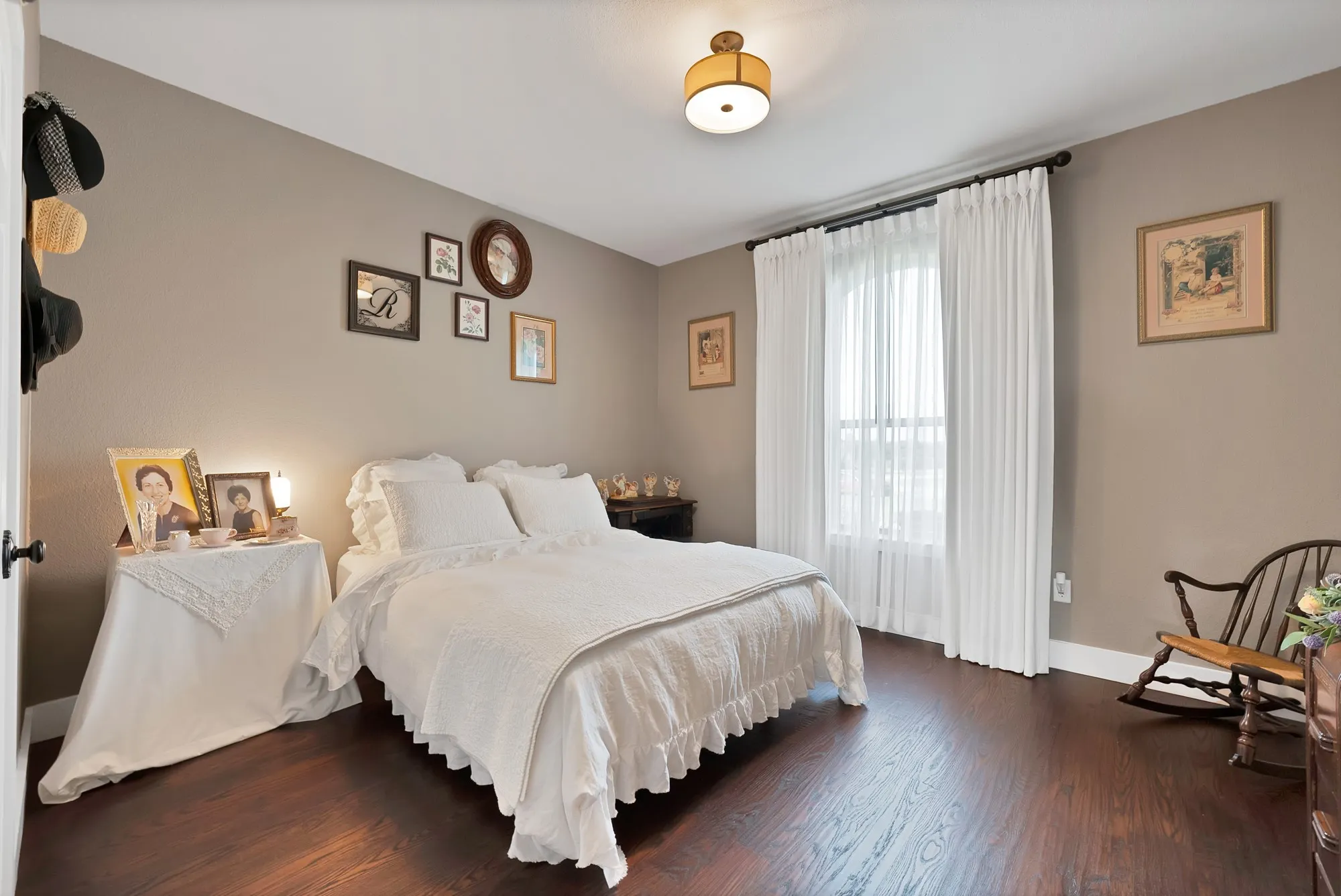 Bedroom featuring rich wood-finish flooring, neutral wall tones, and a window dressed with sheer and opaque curtains