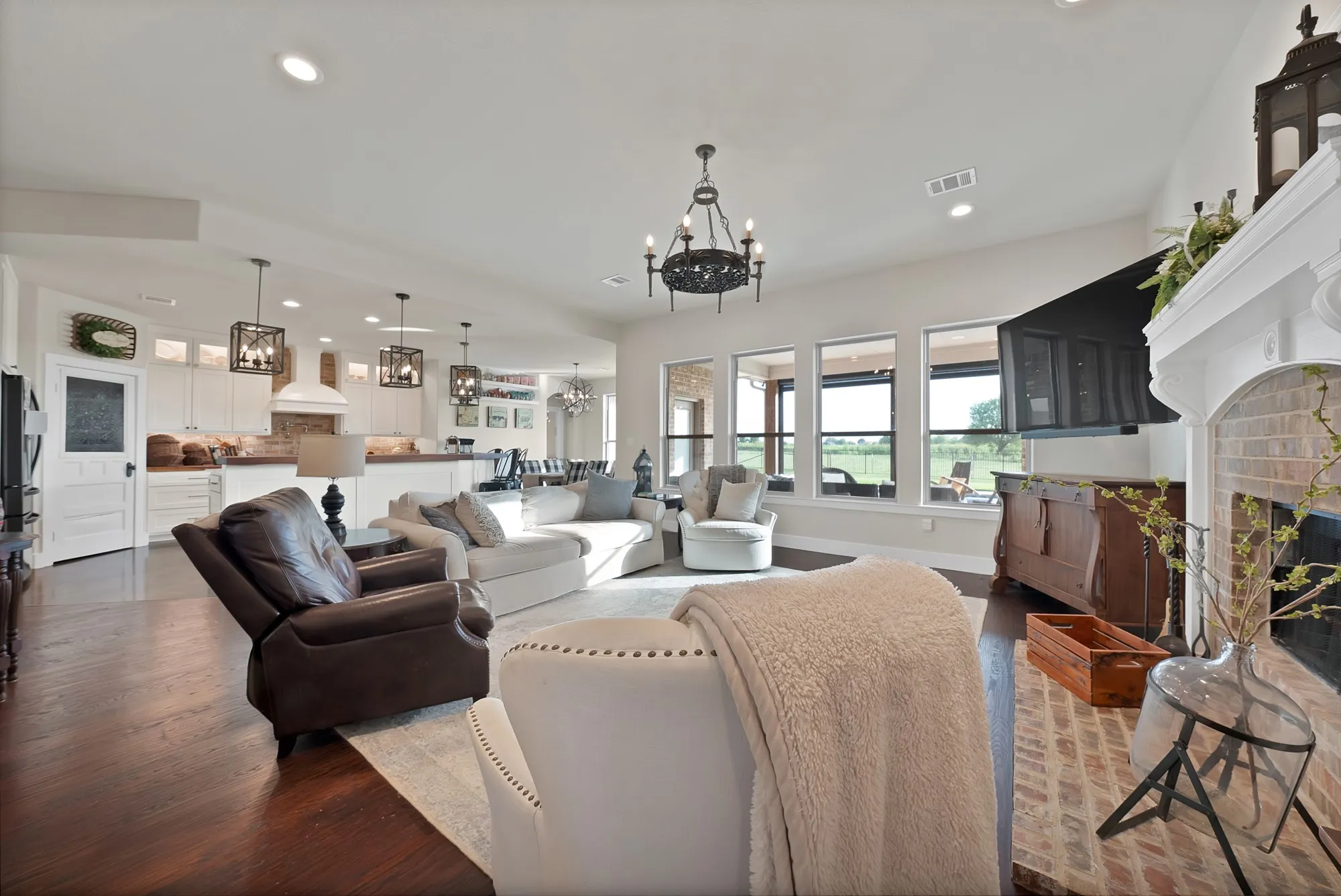 Spacious living area featuring wood-finish flooring, a brick fireplace with a white mantel, and an open-concept design to the kitchen