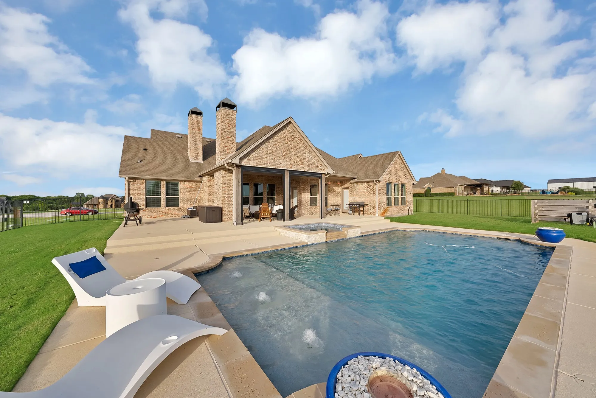 Spacious backyard featuring a large swimming pool with integrated spa, expansive concrete patio, lush green lawn, and a brick exterior home with covered patio