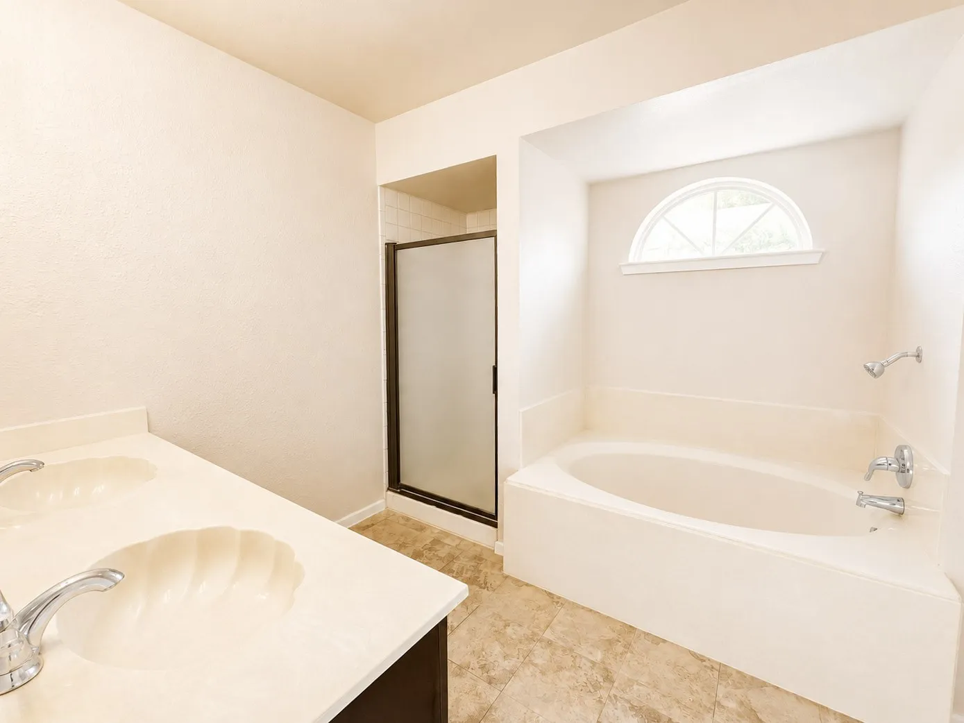 Bathroom featuring a dual vanity with shell-motif sinks, a garden tub, a walk-in shower with a frosted glass door, and an arched transom window