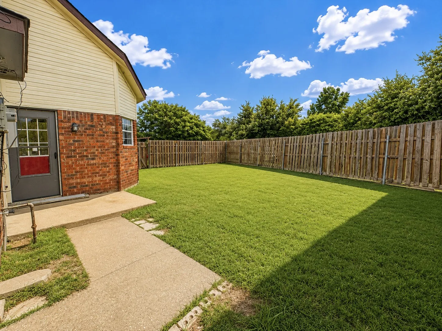 Expansive grassy yard with a wooden privacy fence, concrete patio, and a side entrance with a brick exterior
