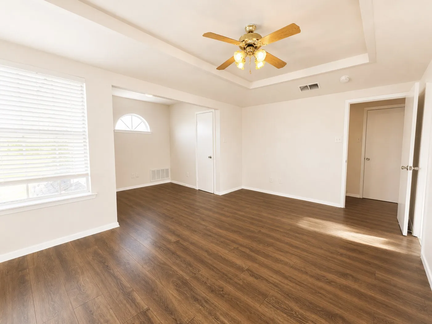Spacious room featuring wood-finish flooring, a tray ceiling with a brass-finish ceiling fan, and white window blinds