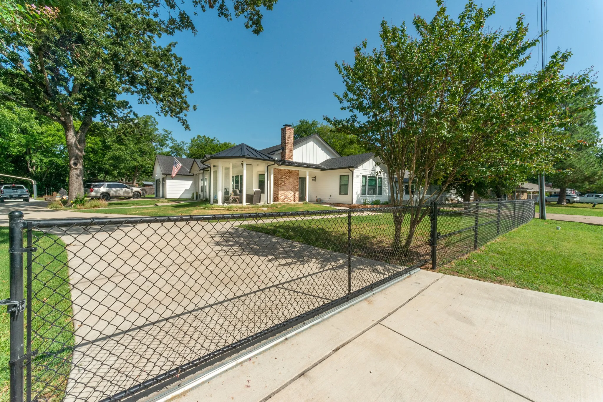 Gated driveway entry leading to a white exterior residence with a brick chimney accent, a black metal roof on a rounded porch, and a side-gabled garage structure