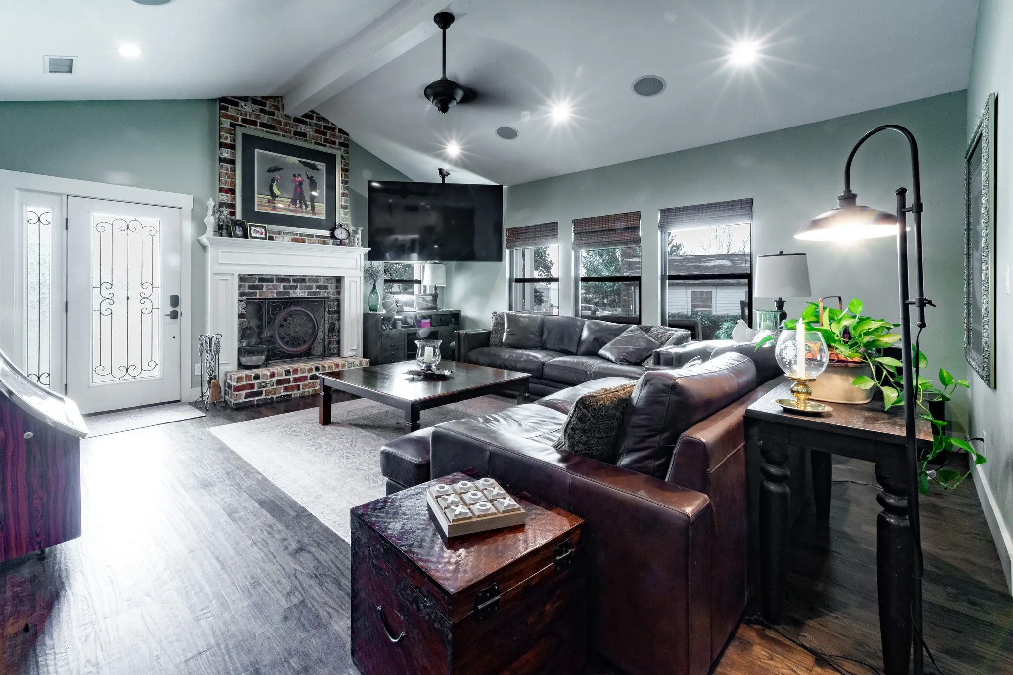 Vaulted ceiling living area featuring wood-finish flooring, a brick-front fireplace with a white mantel, and multiple windows