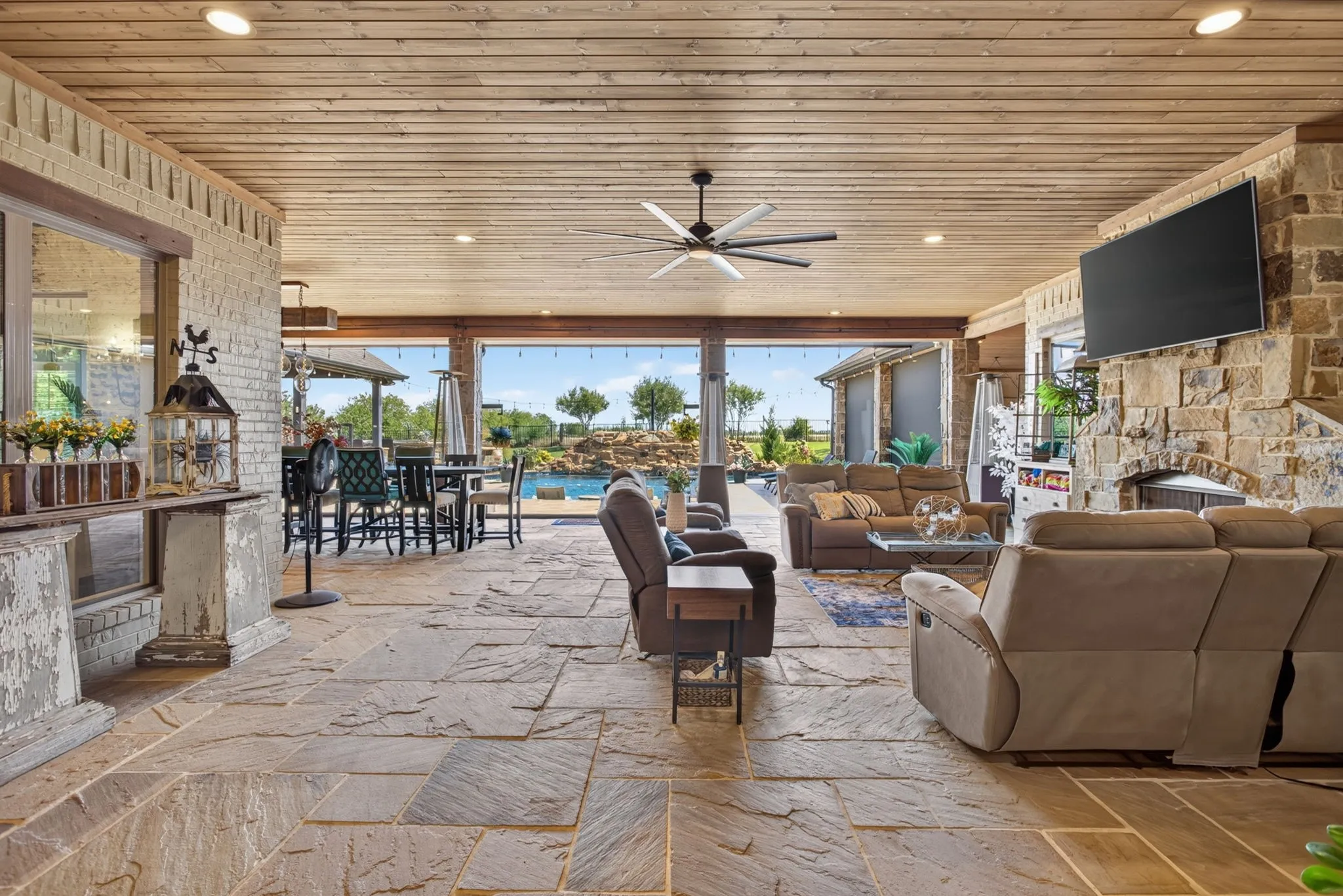 Expansive covered patio featuring a flagstone floor, wood-plank ceiling with recessed lighting and a ceiling fan, stone-clad fireplace, and an outdoor dining area with views of the swimming pool