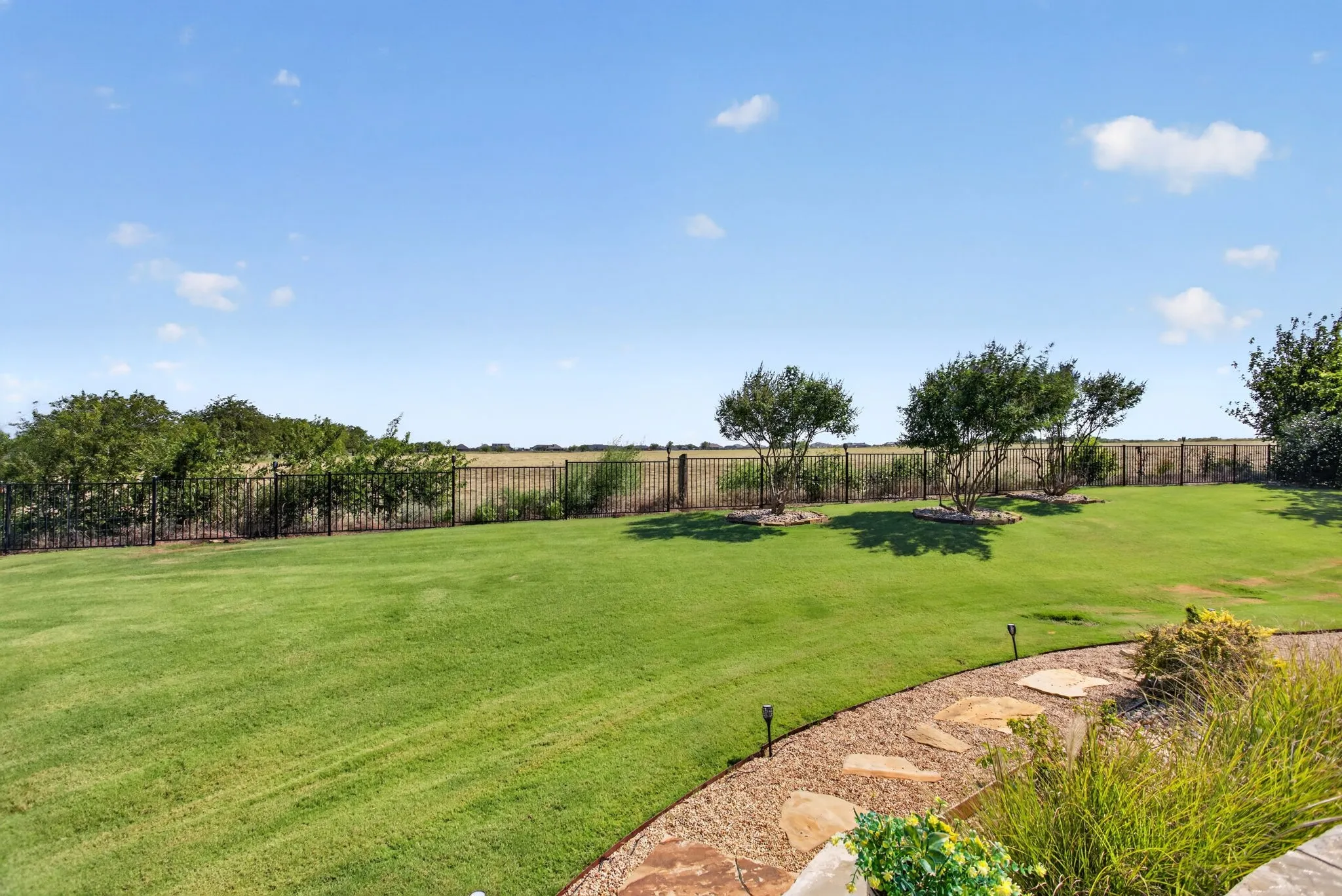 Expansive lawn area featuring a black metal fence, mature trees, and a gravel pathway with natural stone accents