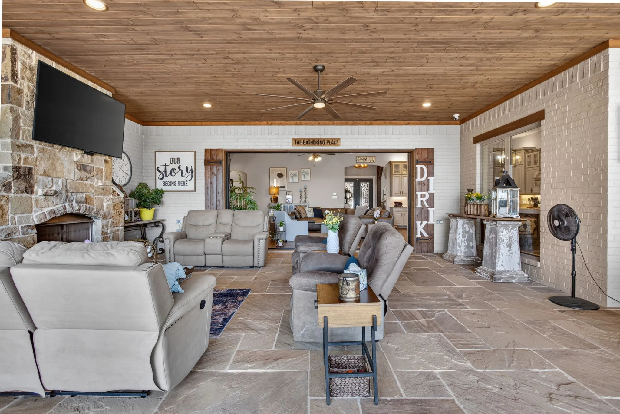 Expansive covered patio featuring a stone-clad fireplace, wood-plank ceiling with recessed lighting, large format stone flooring, and white brick walls