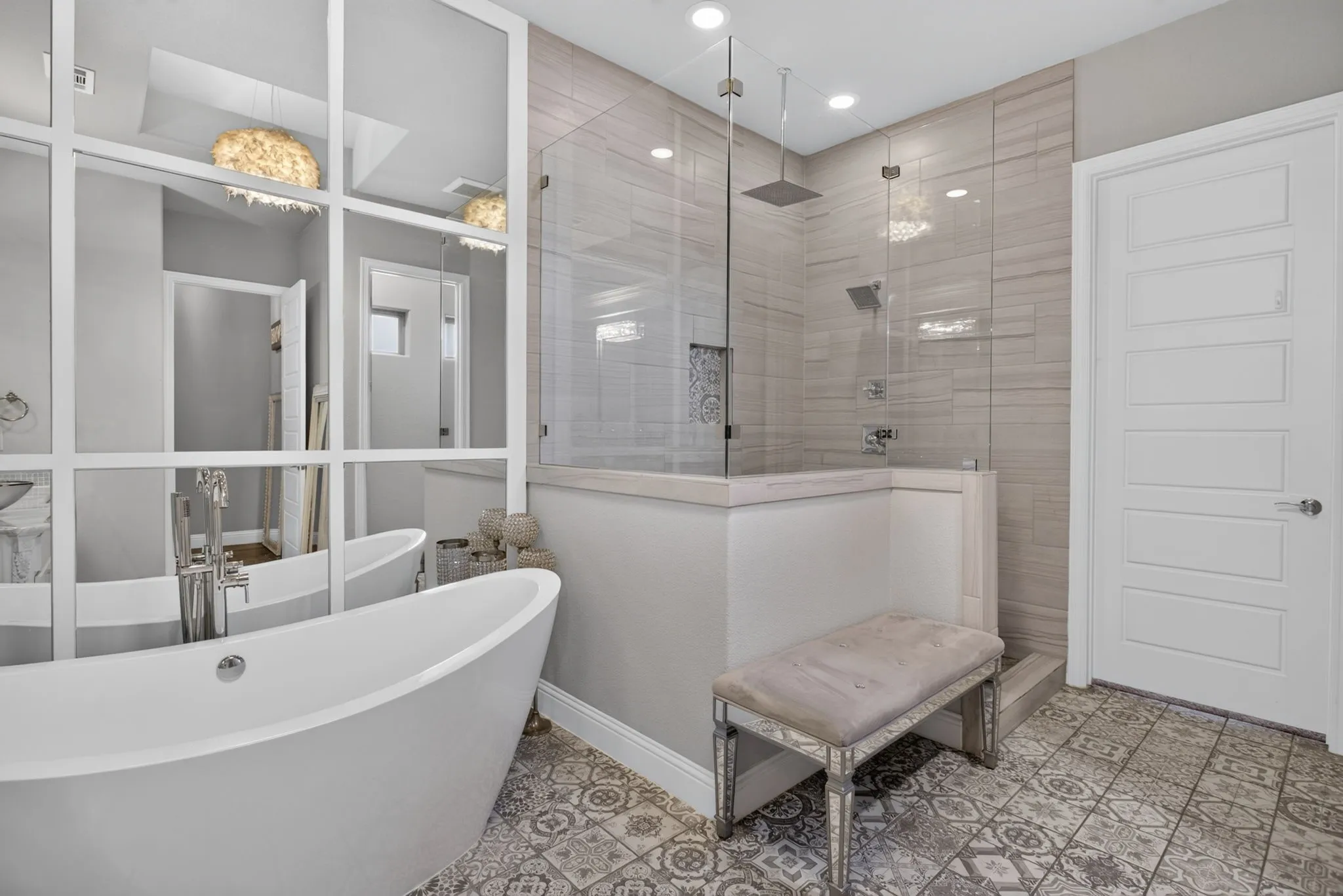Freestanding soaking tub with floor-mounted faucet, glass-enclosed shower with rain head, patterned floor tiling, tiled shower walls, and a multi-panel interior door