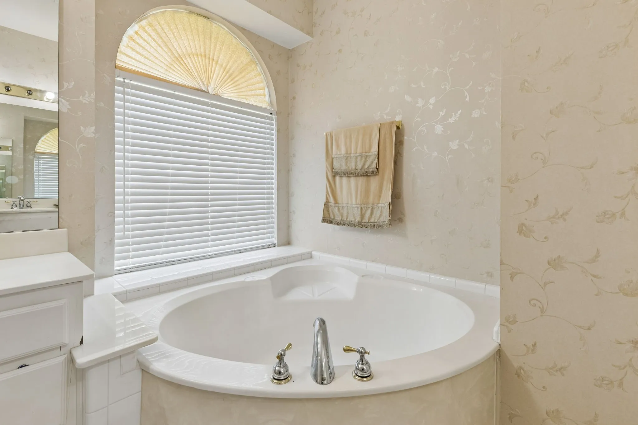 Corner soaking tub with a tiled surround, arched window featuring horizontal blinds and a pleated shade, vanity cabinet with a countertop, and decorative wallpaper
