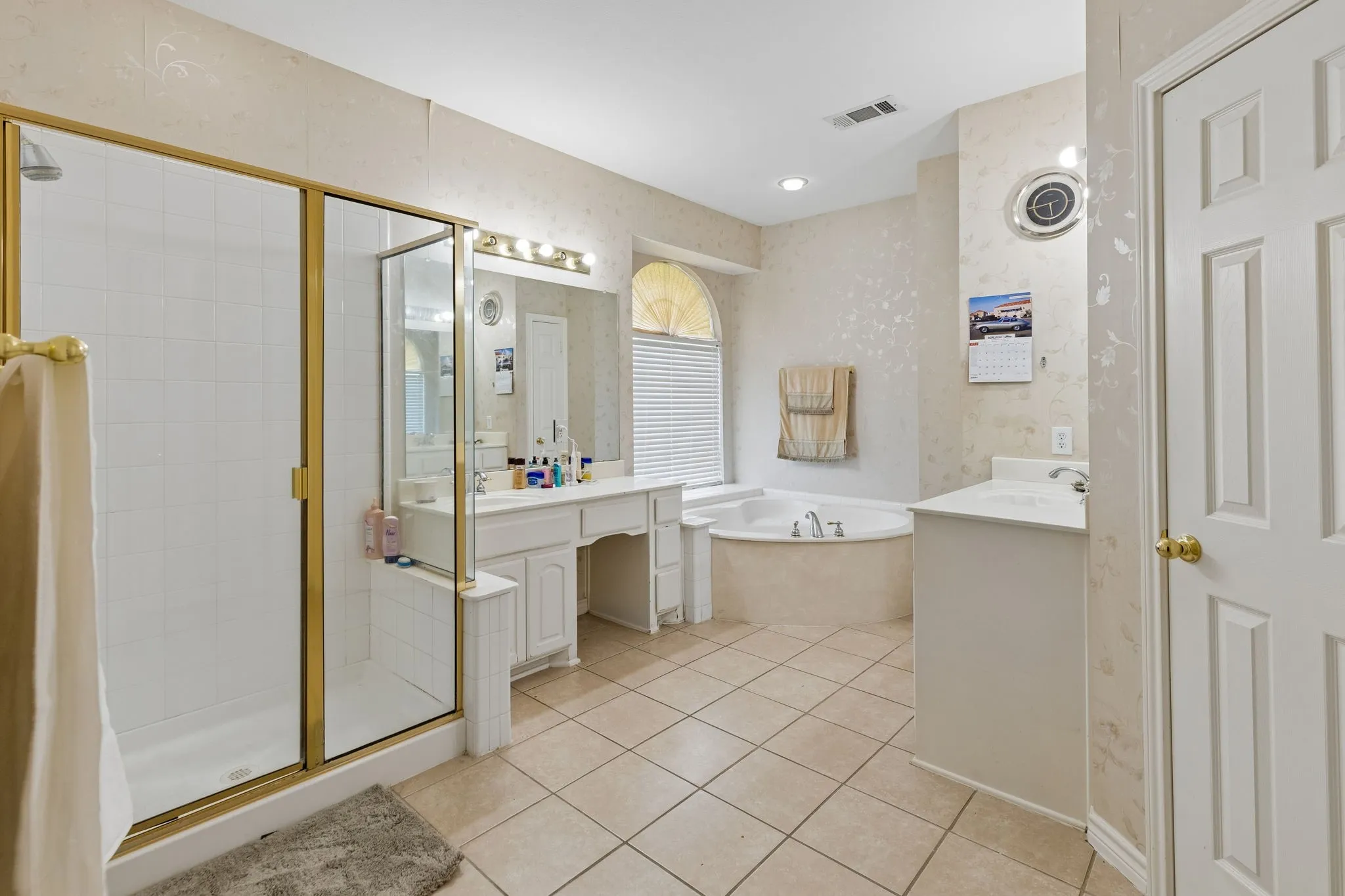 Ceramic tile flooring in an expansive bathroom featuring a glass-enclosed shower with gold-tone framing, a jetted soaking tub, a double vanity with a makeup area, and a single vanity