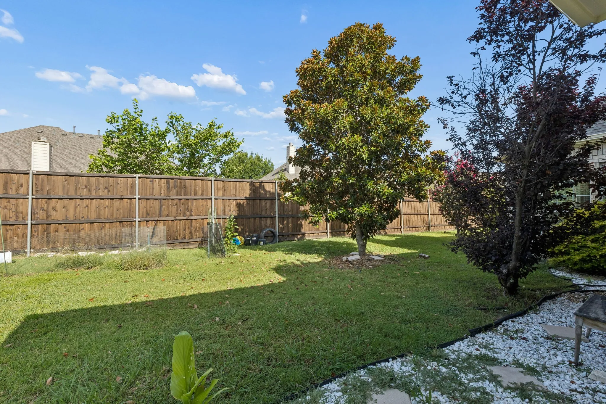 Fenced backyard featuring mature trees, a manicured lawn, and a decorative white stone border