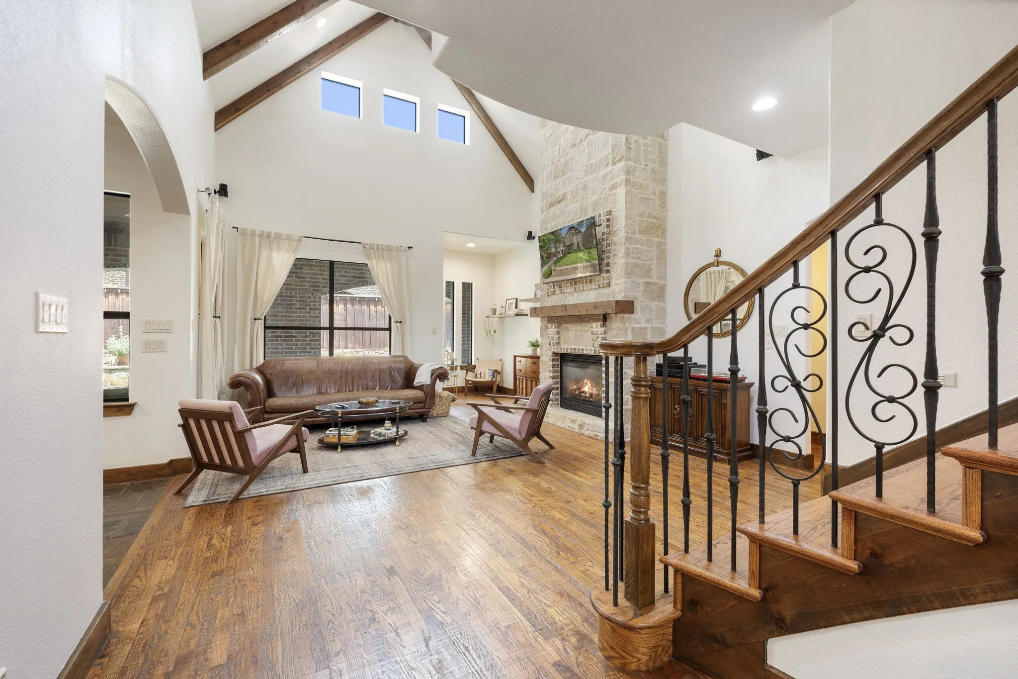 Vaulted ceiling with exposed wood beams, stone-clad fireplace, wood-finish flooring, staircase with wrought iron balusters, and arched entryway