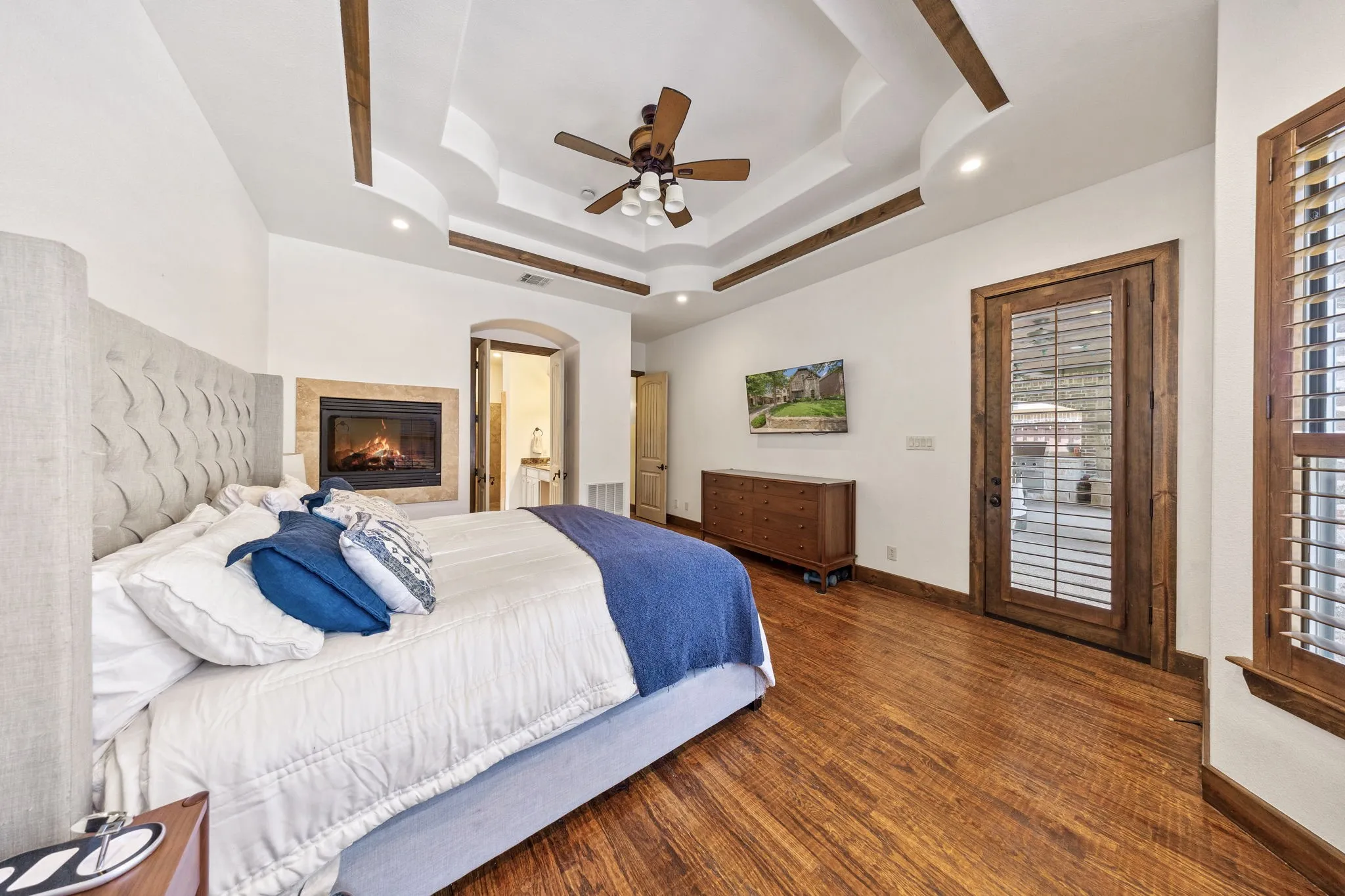 Master bedroom featuring a coffered ceiling with recessed lighting and ceiling fan, wood-finish flooring, a gas fireplace, and French doors with plantation shutters opening to natural light