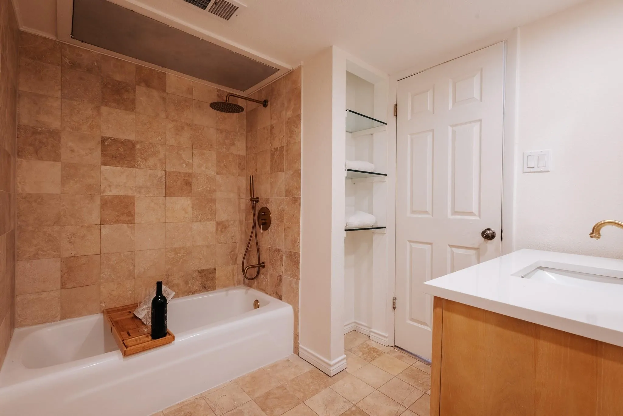 Bathroom featuring a tiled shower and tub combination with a rain shower head, a wood-finish vanity with an integrated sink and white countertop, a white panel door, and built-in glass shelving