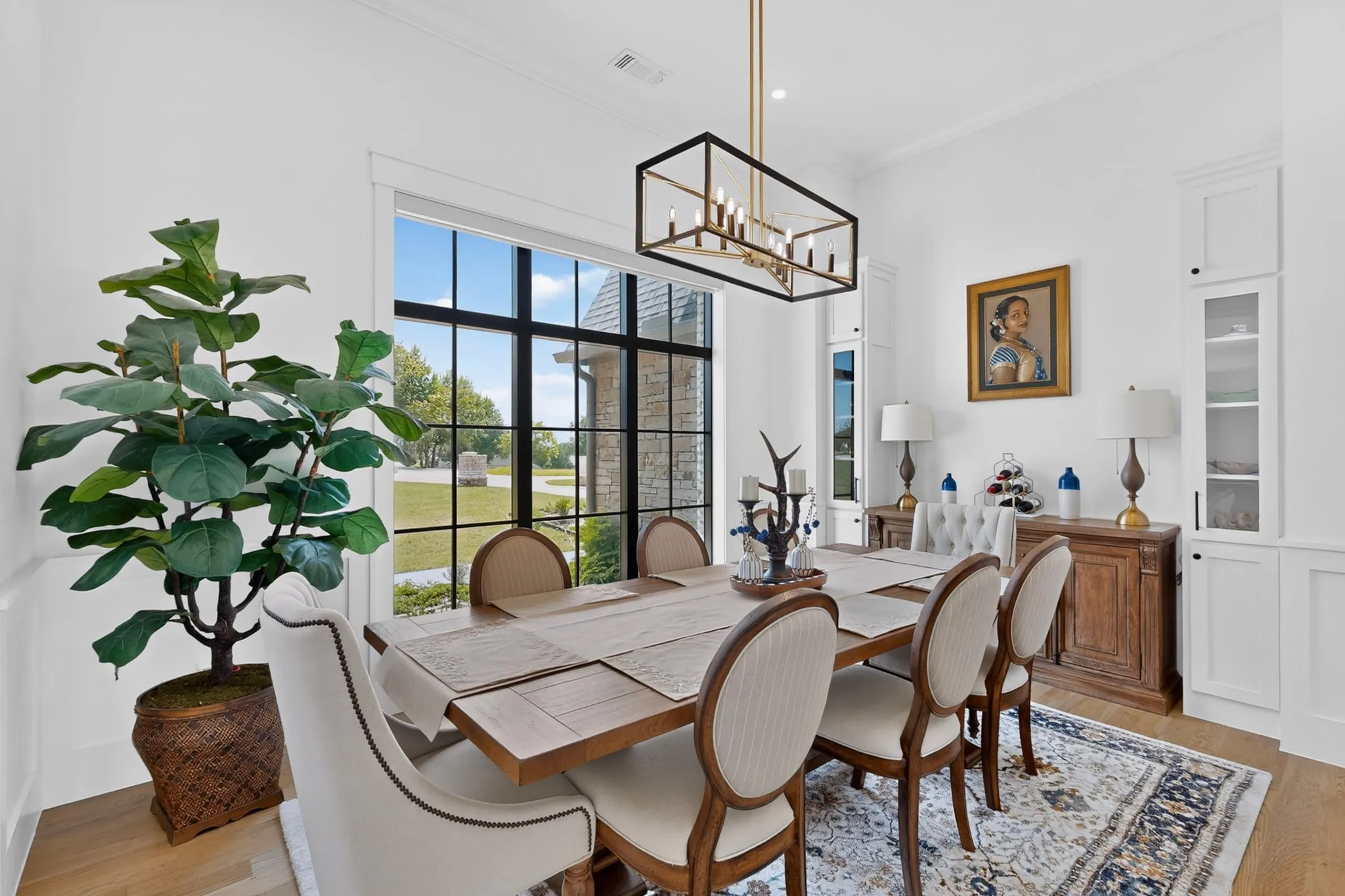 Dining area featuring a modern linear chandelier, large picture window with grid detailing, light wood-finish flooring, and built-in cabinetry with glass display panels